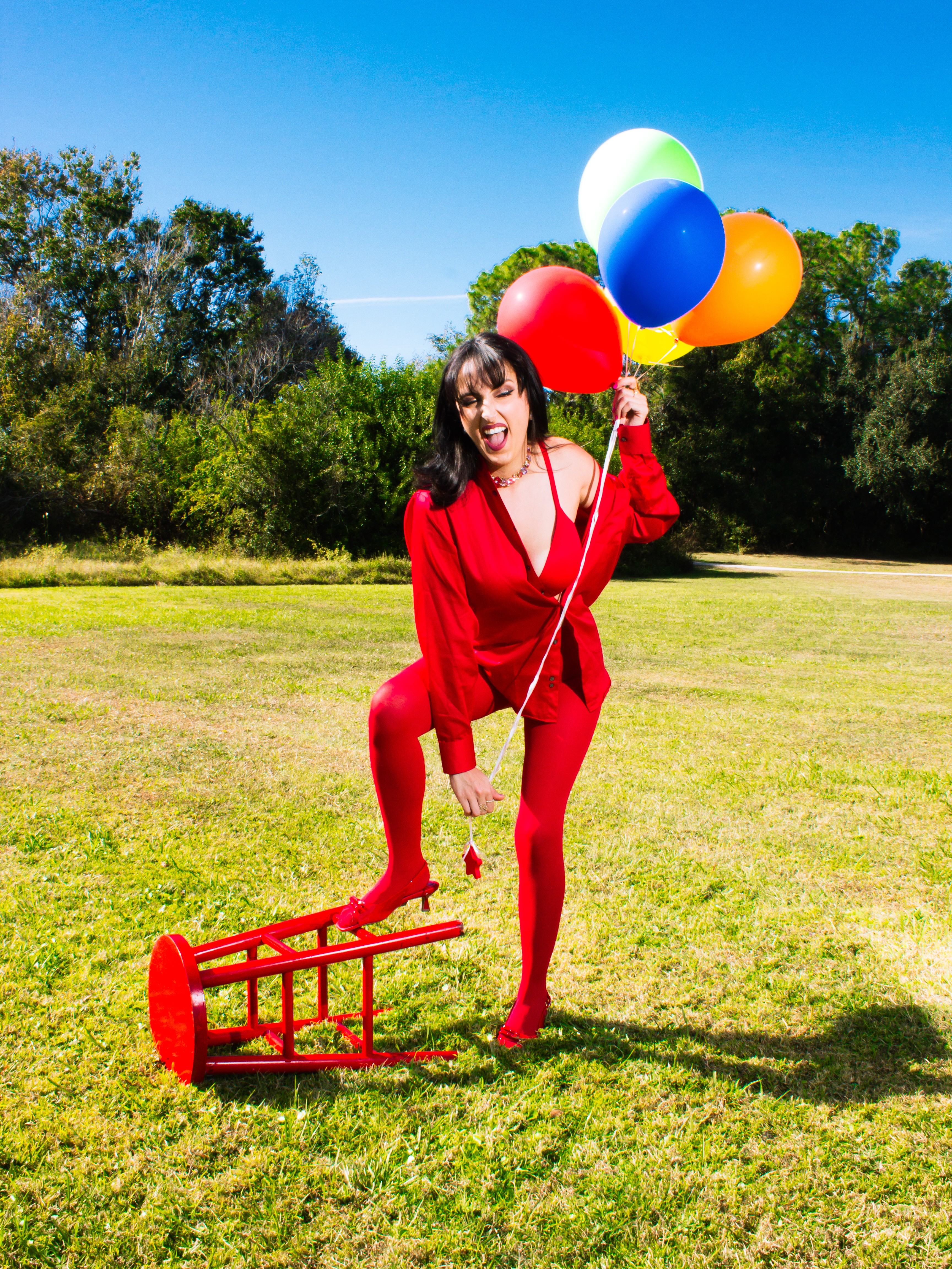 Woman in red outfit holding balloons and a small red rake in a grassy field with trees and blue sky.