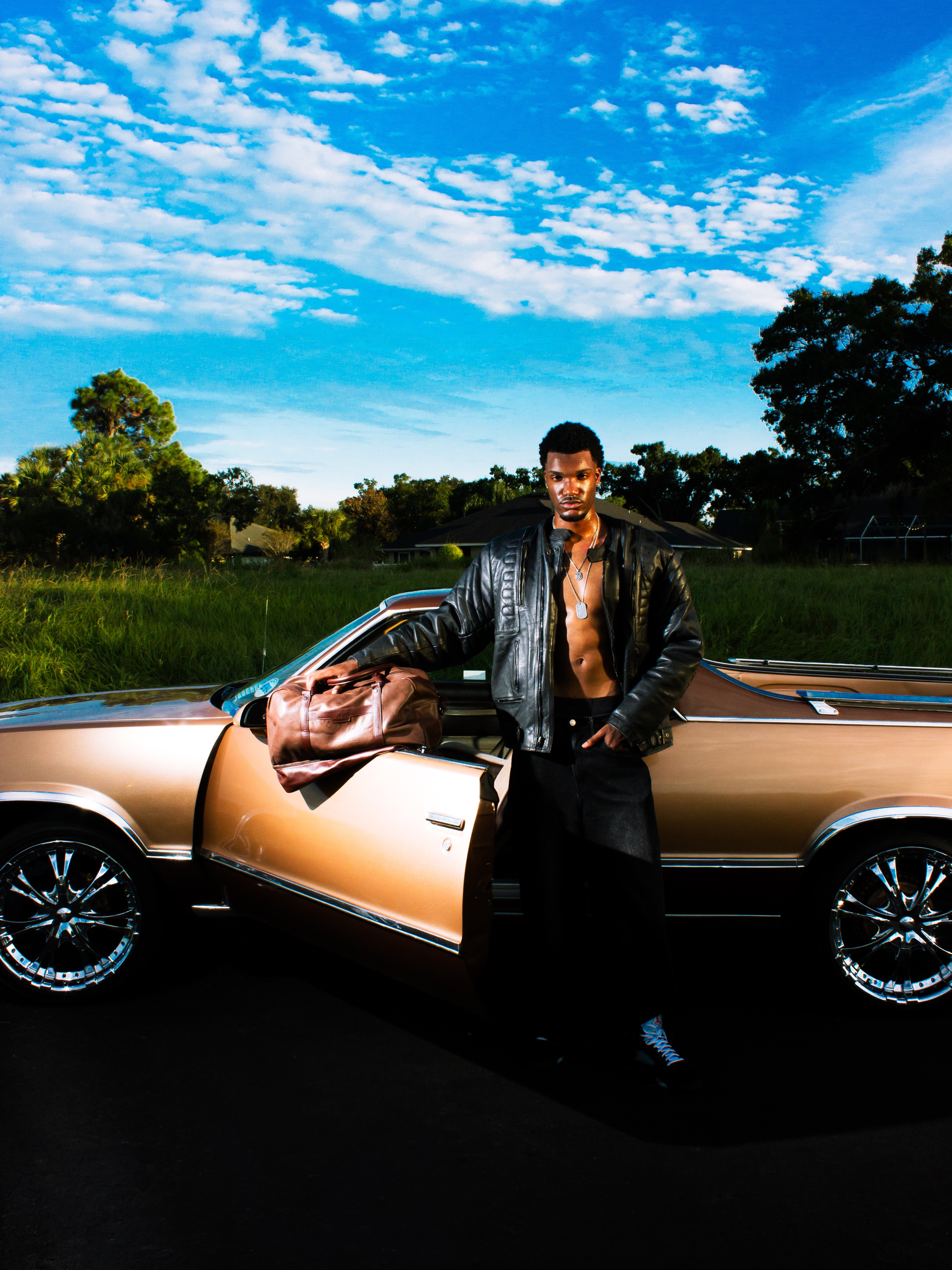 Man standing next to a vintage car outdoors under a blue sky with clouds.