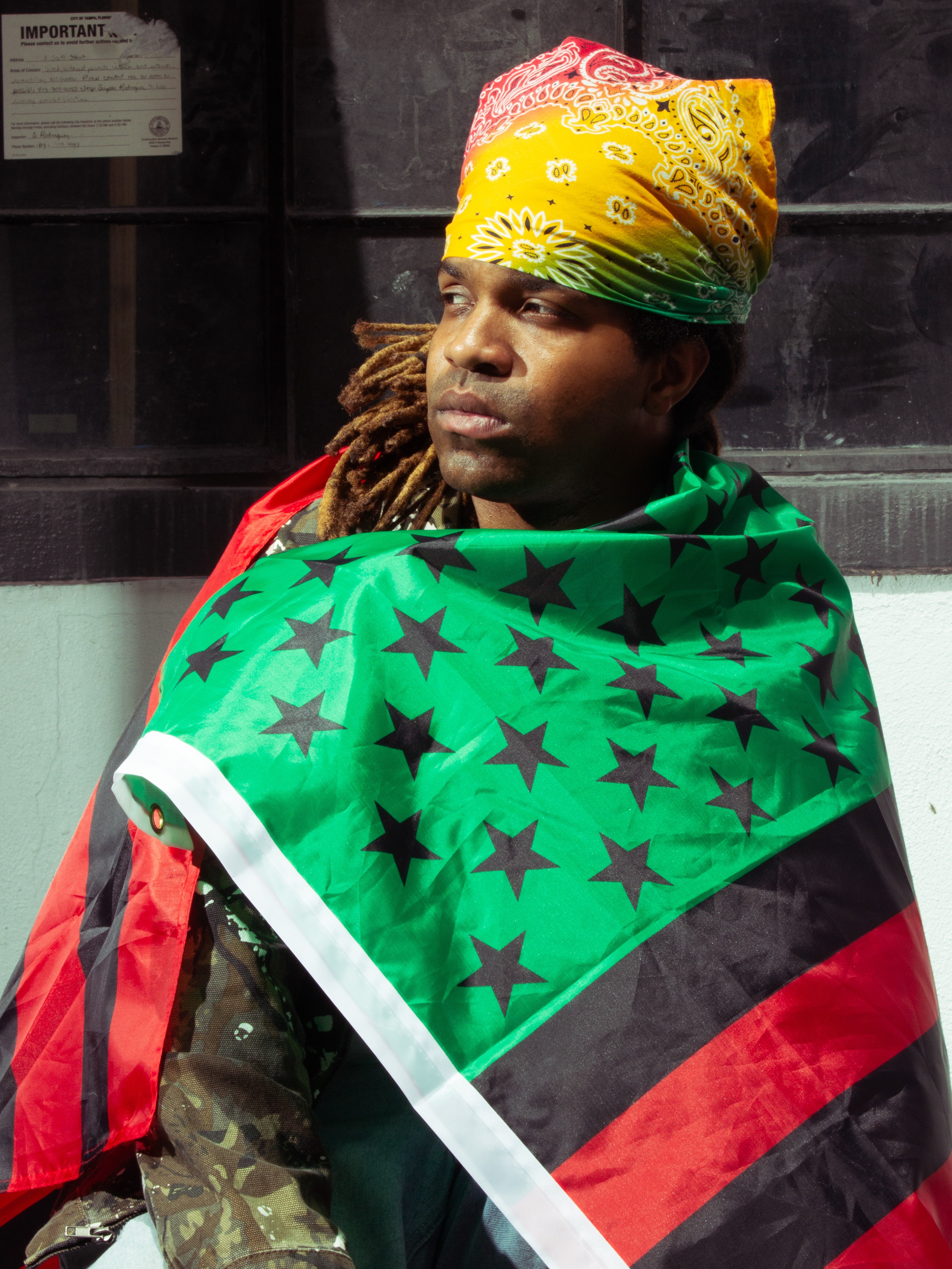 Person with colorful headwrap and green star-patterned flag draped over shoulders, looking to the side indoors.