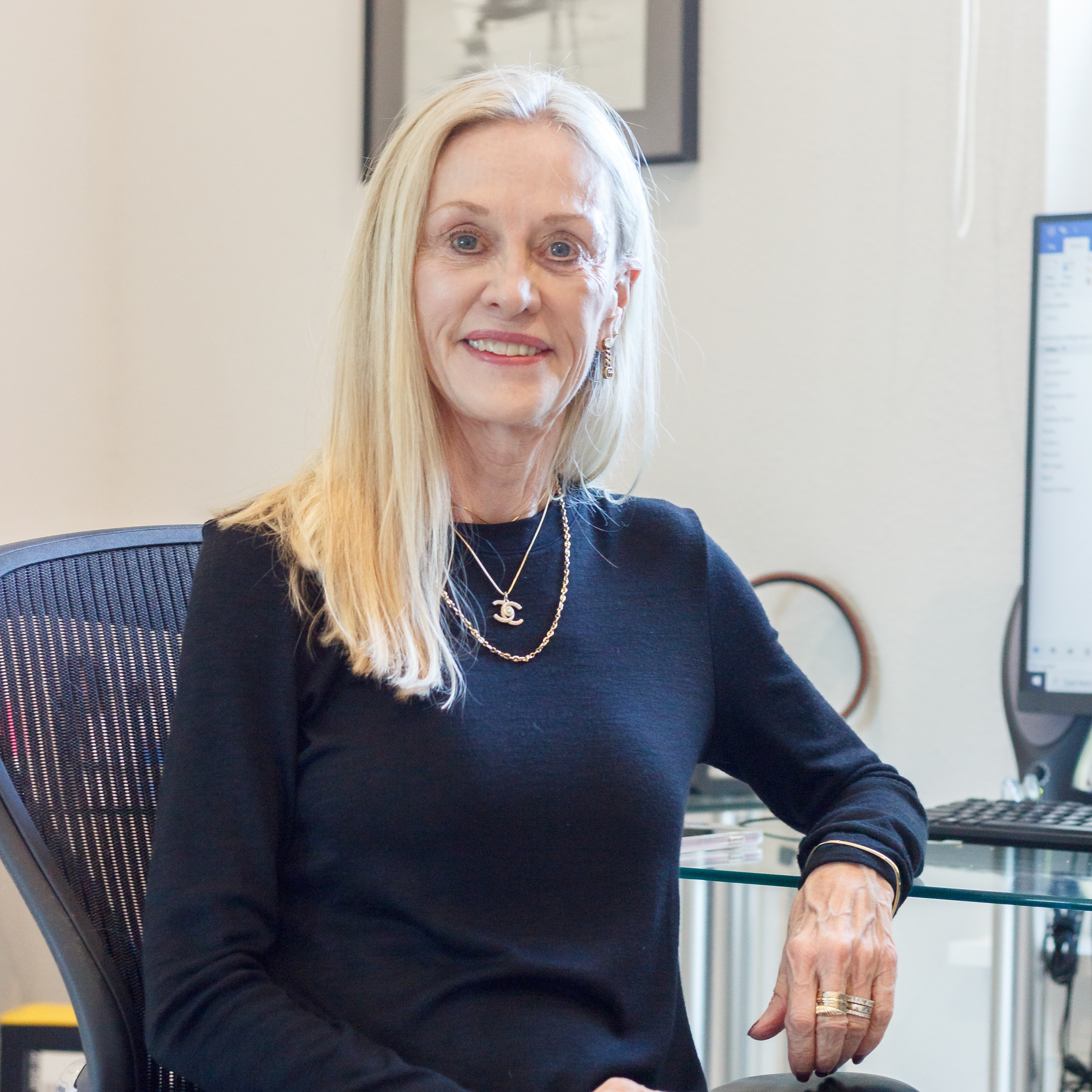 Woman with long blonde hair sitting at desk, smiling, in an office with computer monitor and framed picture.