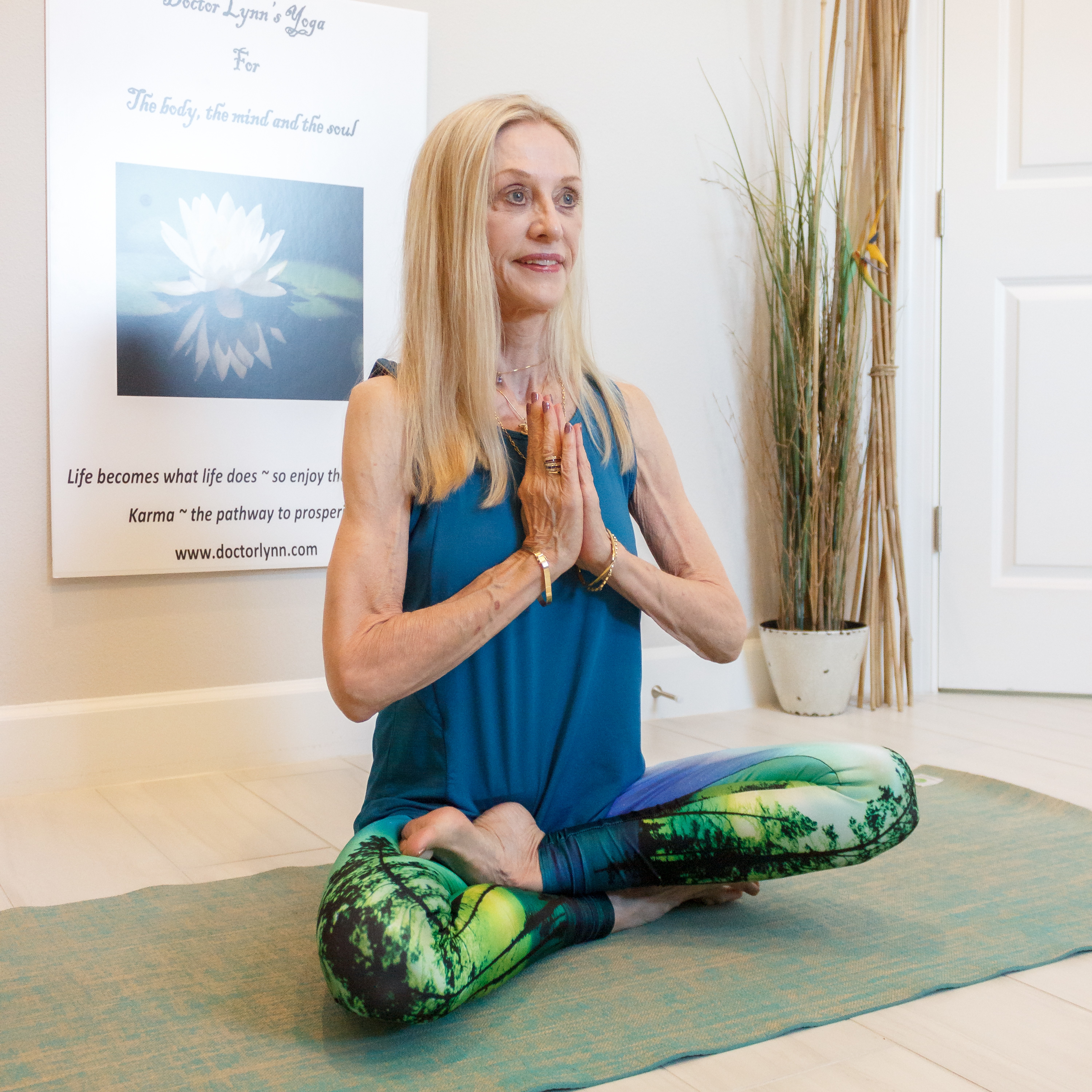 Woman practicing yoga in a room with a poster and a plant, sitting cross-legged with hands in prayer position.