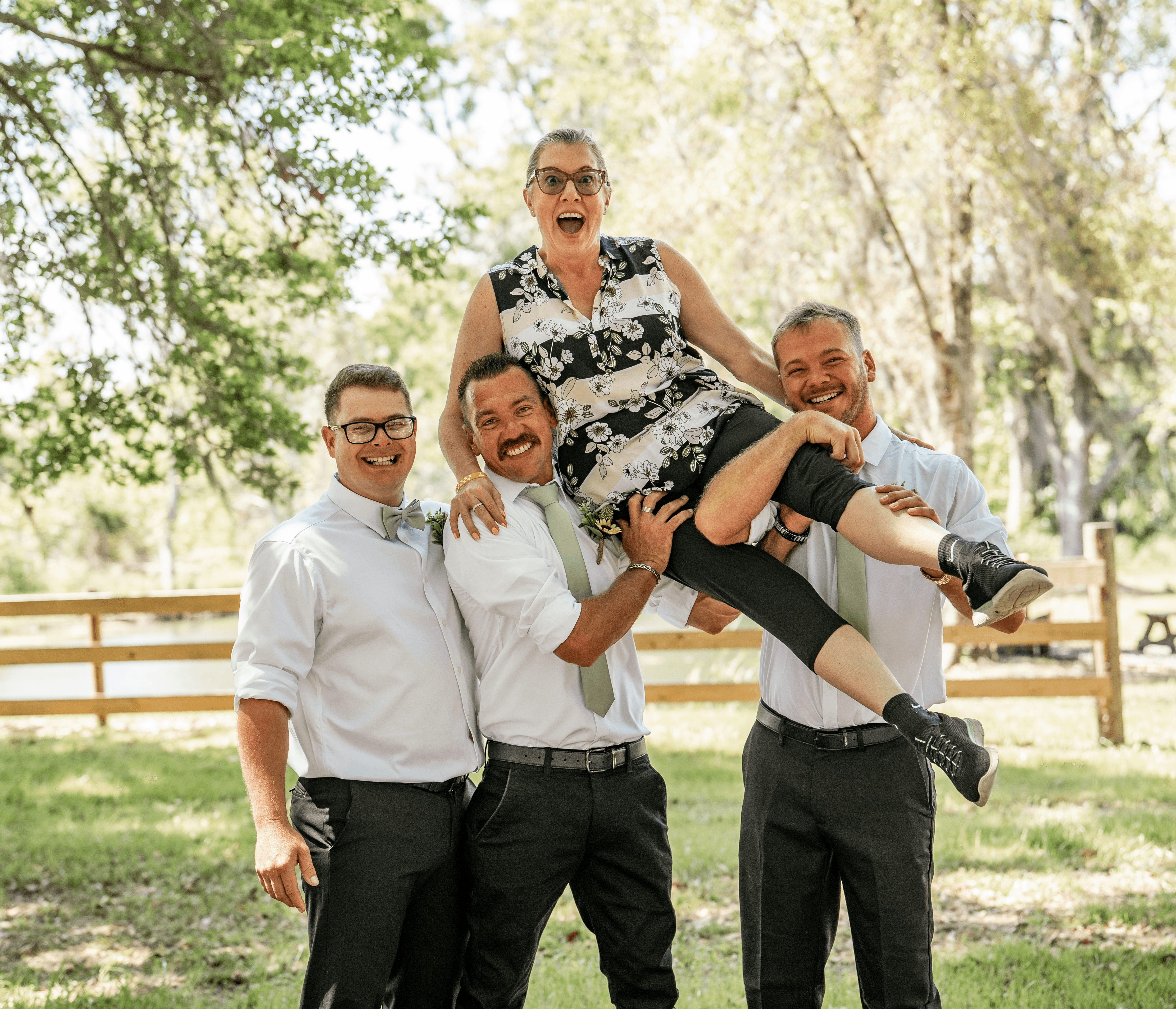 Four people outdoors, three men standing, woman being lifted, all smiling, trees and fence in background.