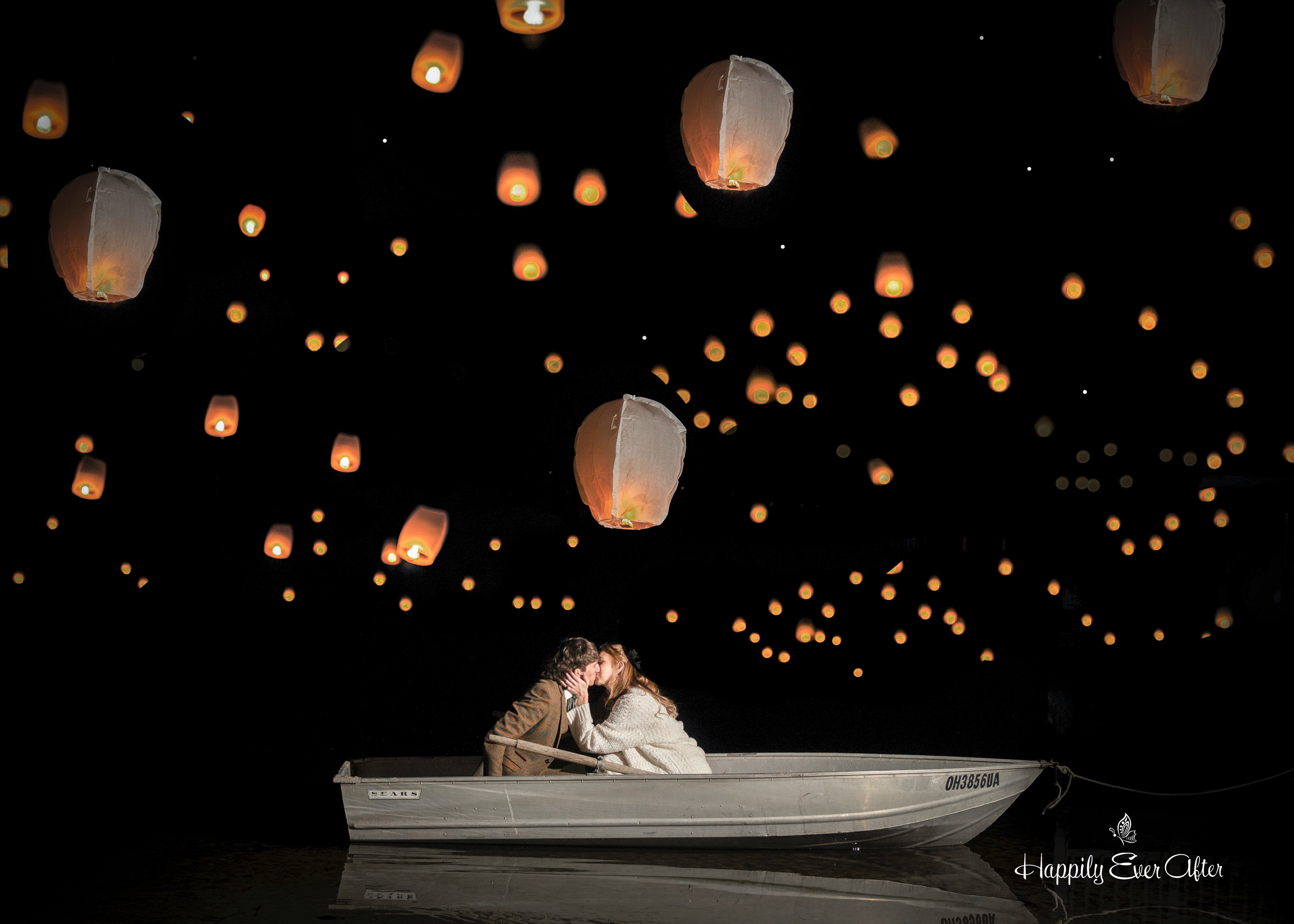 Person sitting in a boat at night with floating lanterns in the sky above.
