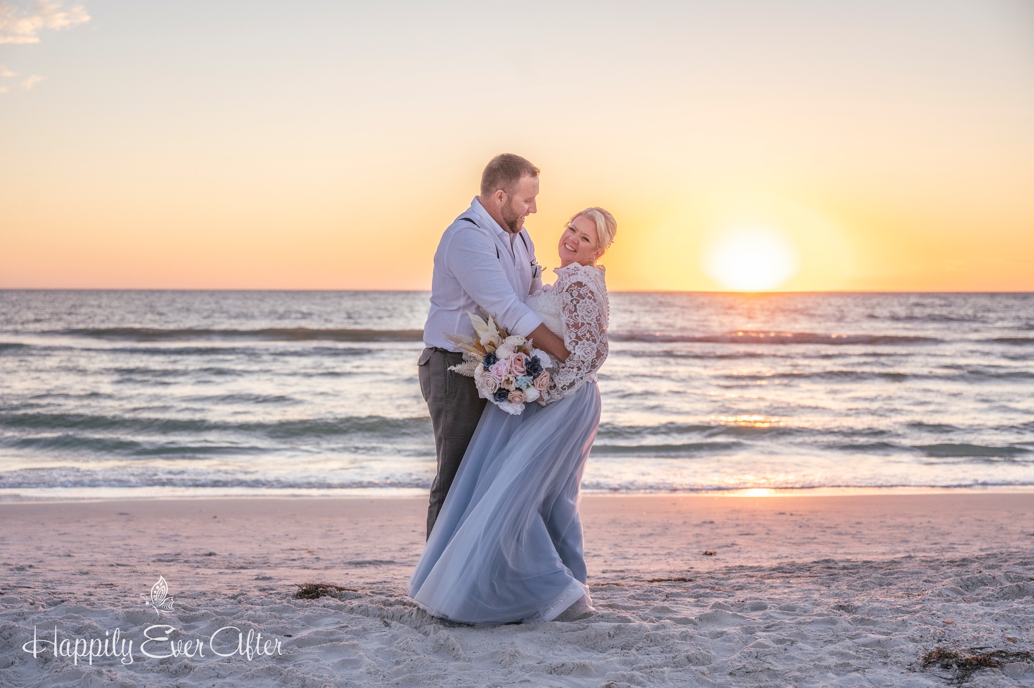 Couple in wedding attire standing on beach at sunset, embracing and smiling.