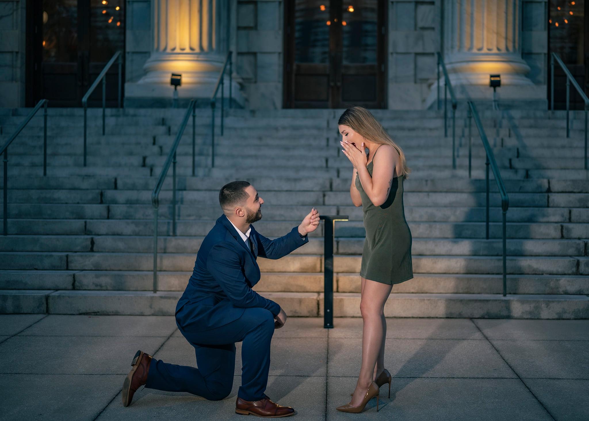 Man kneeling proposing to woman on steps outside building at night, woman covering face in surprise.