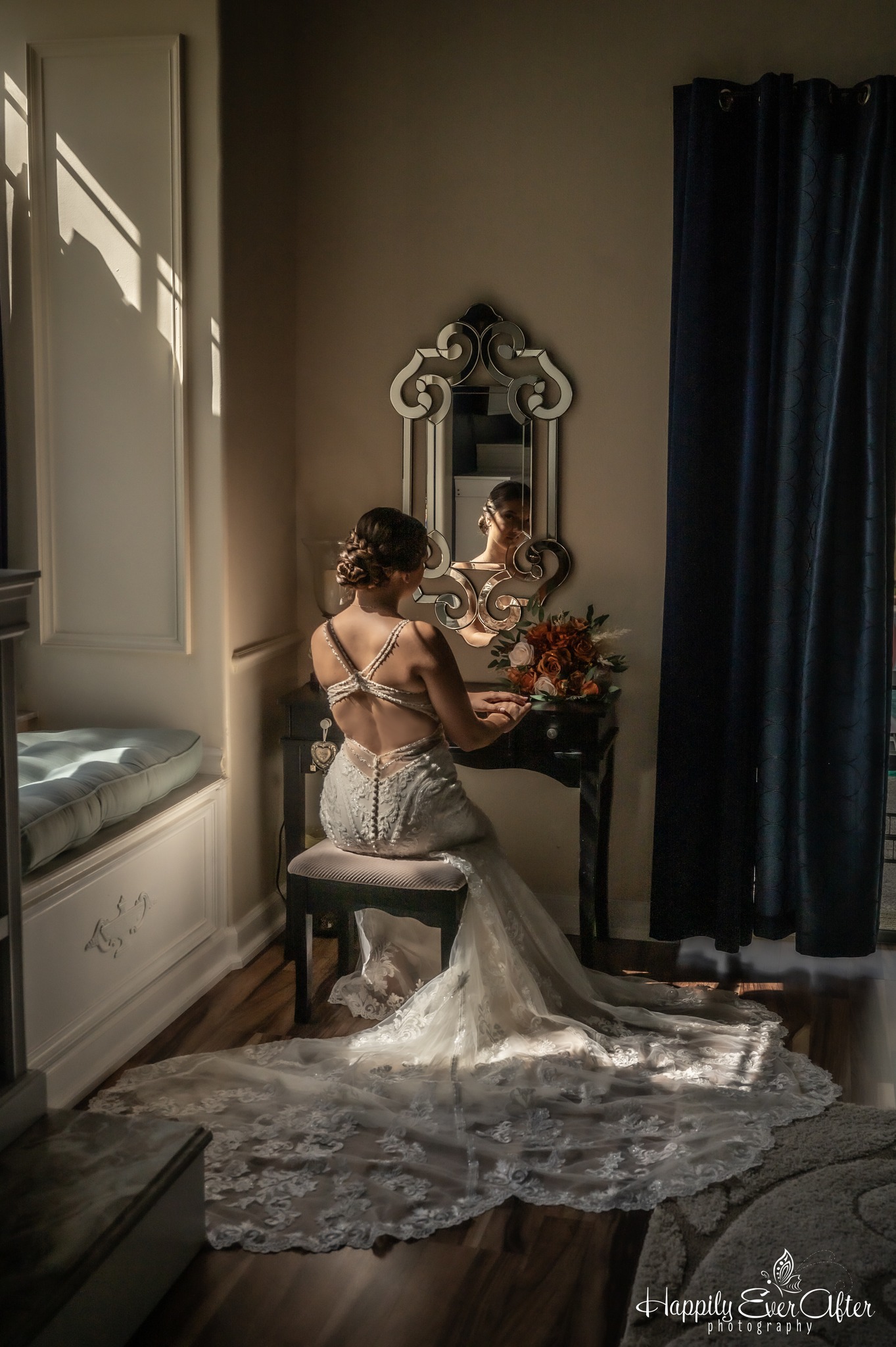 Woman in wedding dress sitting on stool in front of mirror with flowers on table, sunlight through window, dark curtains nearby.