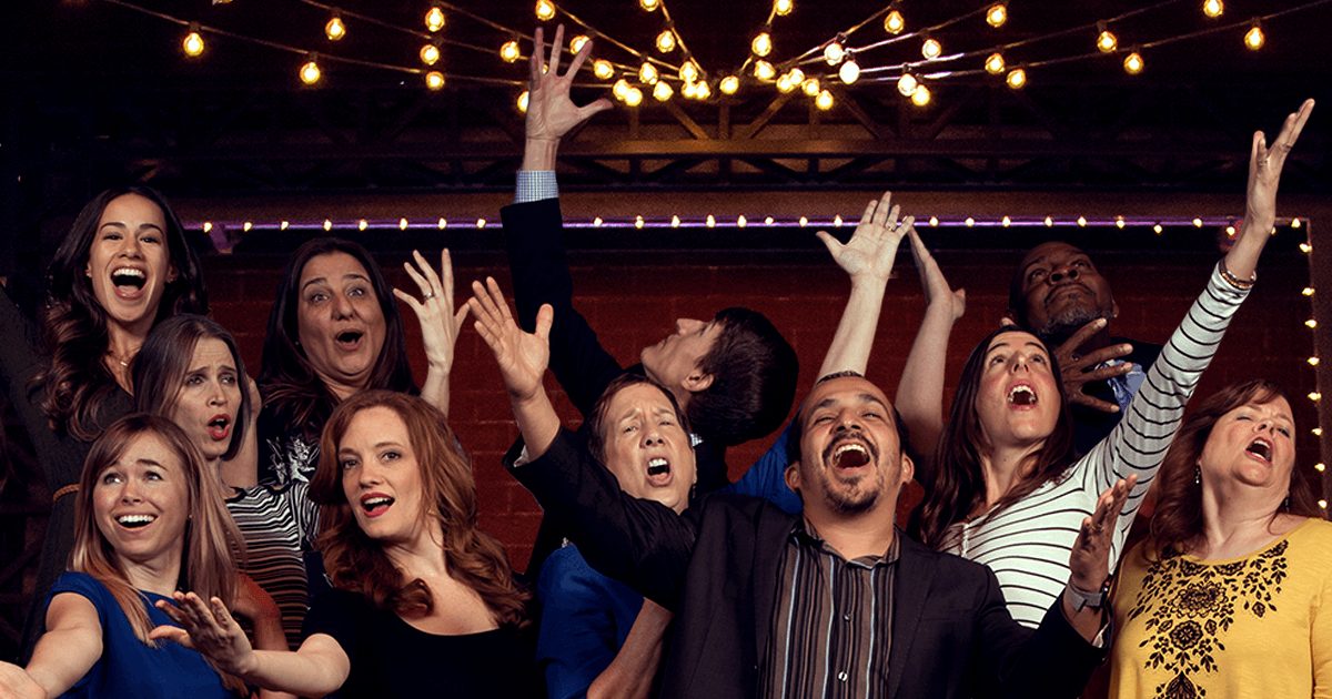 Group of people celebrating indoors with raised hands and smiling faces, illuminated by string lights overhead.
