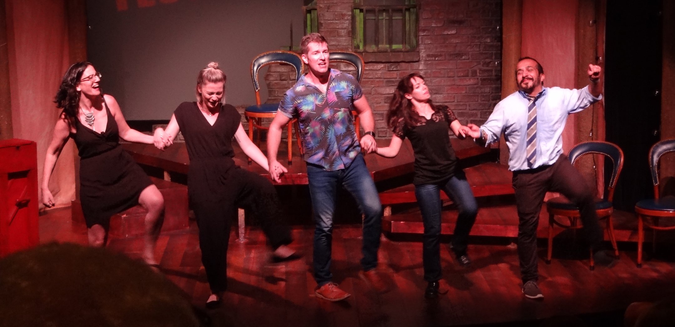 Five people holding hands and dancing on stage with a brick wall background.