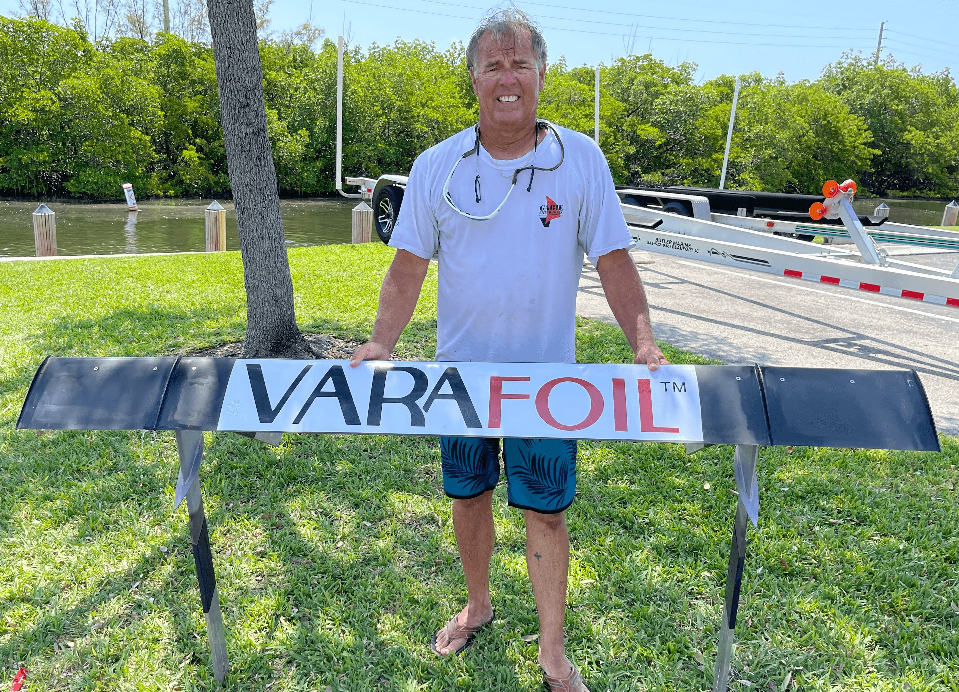 Man standing outdoors behind a large black and white sign reading VARA FOIL, with trees and a boat ramp in background.