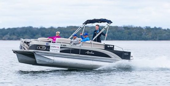 People on a pontoon boat with a canopy, sailing on a lake with a distant shoreline and trees.