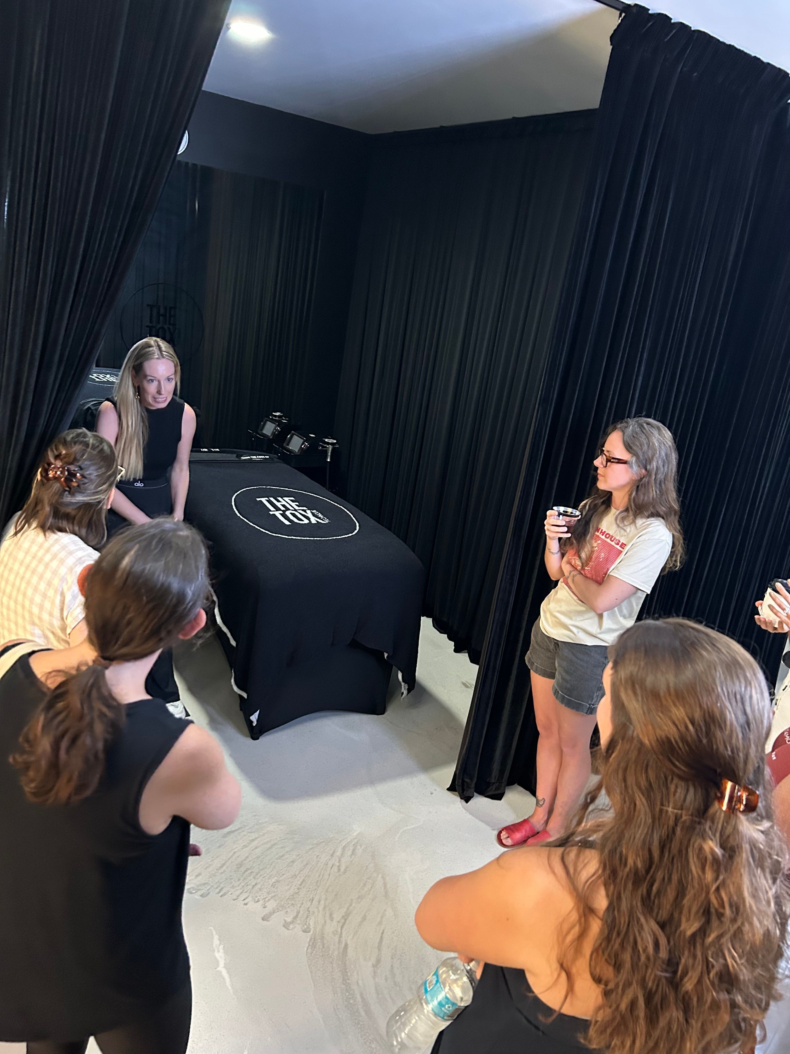 Group of women gathered around a table with a black cloth, one woman speaking into a microphone, others listening.