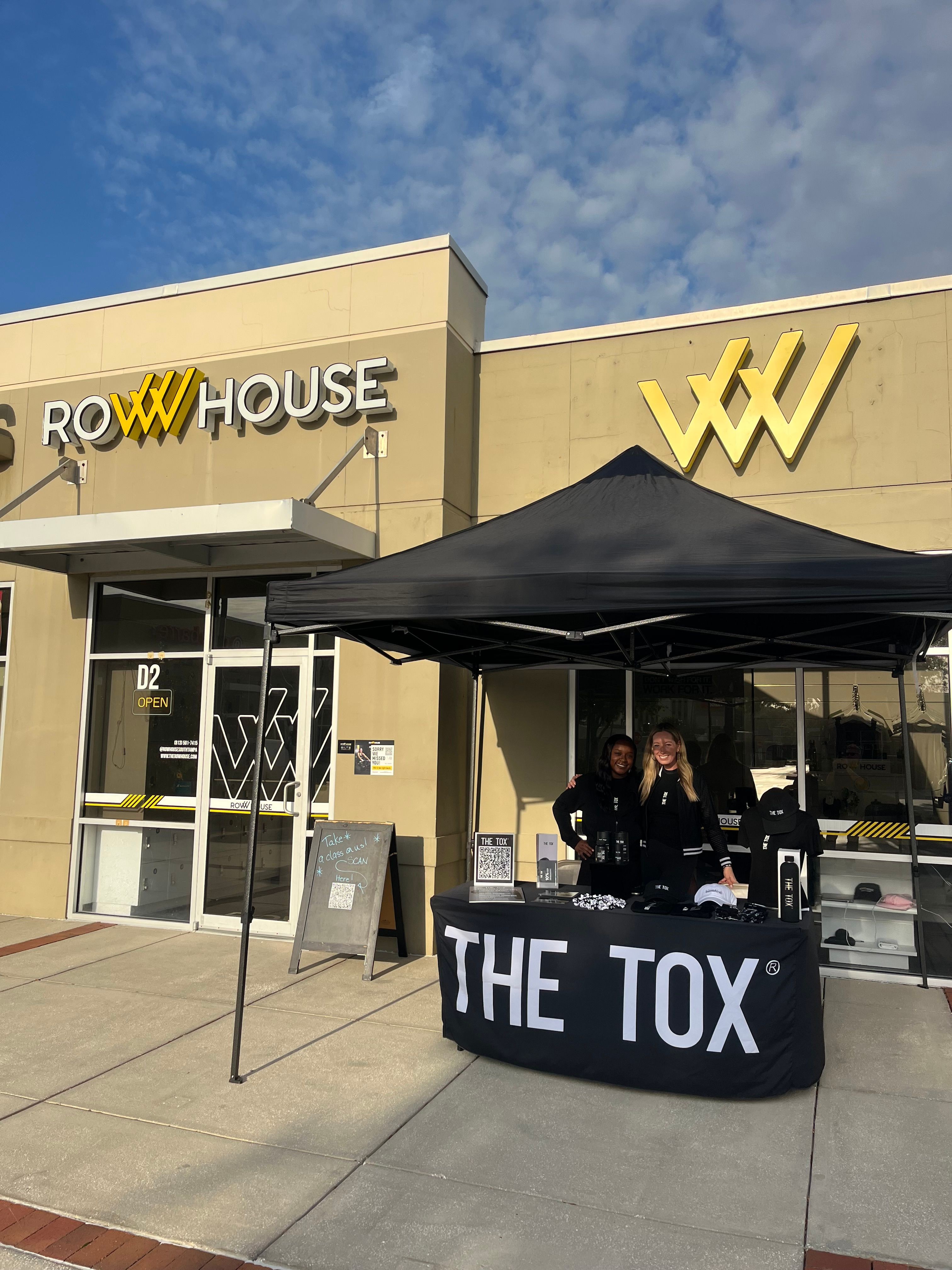 Outdoor booth with two people behind a table under a black canopy in front of a building with a yellow logo.