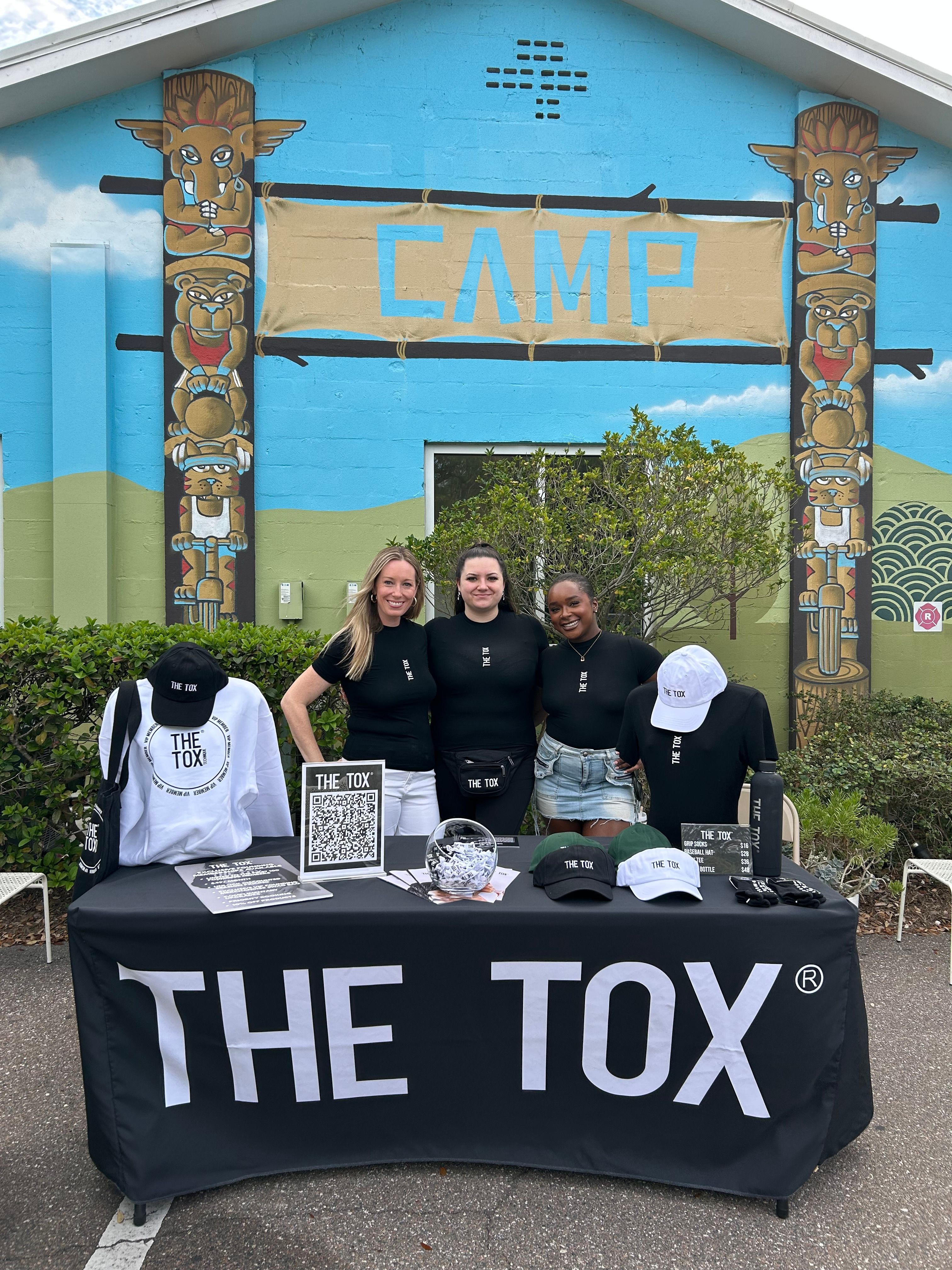 Three women standing behind a table with promotional items at a camp entrance with colorful totem poles and a blue sign.