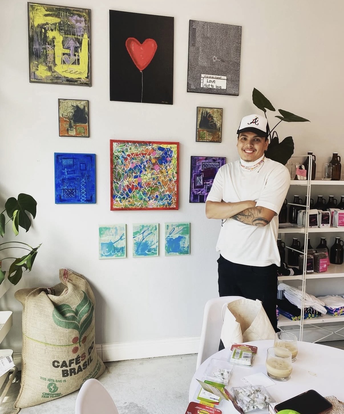 Young person with arms crossed, smiling, standing in art gallery with colorful paintings on white wall, plants, and shelves.