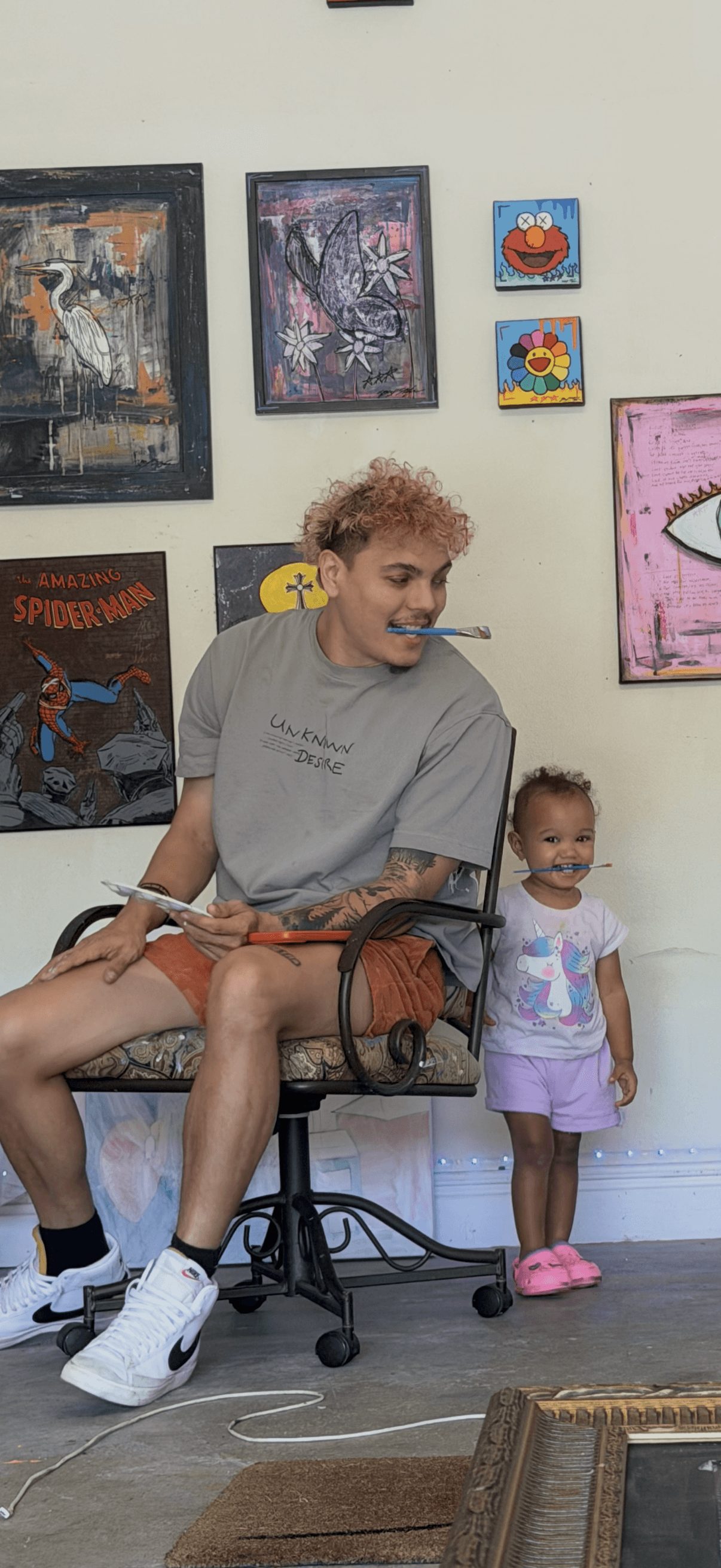 Young man with curly hair sitting on a chair, smiling at a small girl standing nearby in a room with colorful artwork.