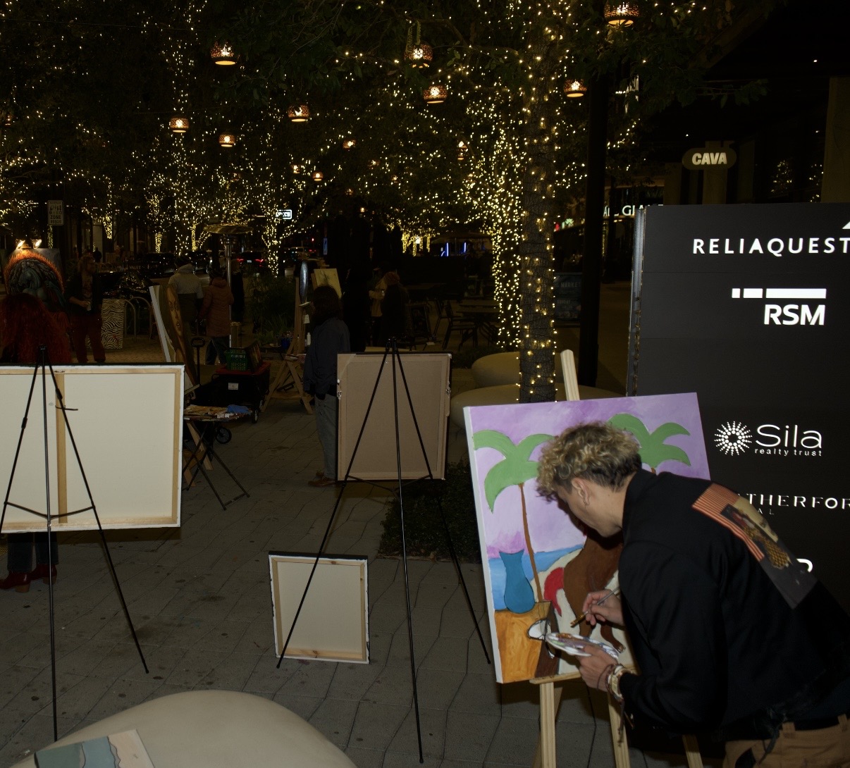 Person painting on canvas outdoors at night with string lights and trees in background.