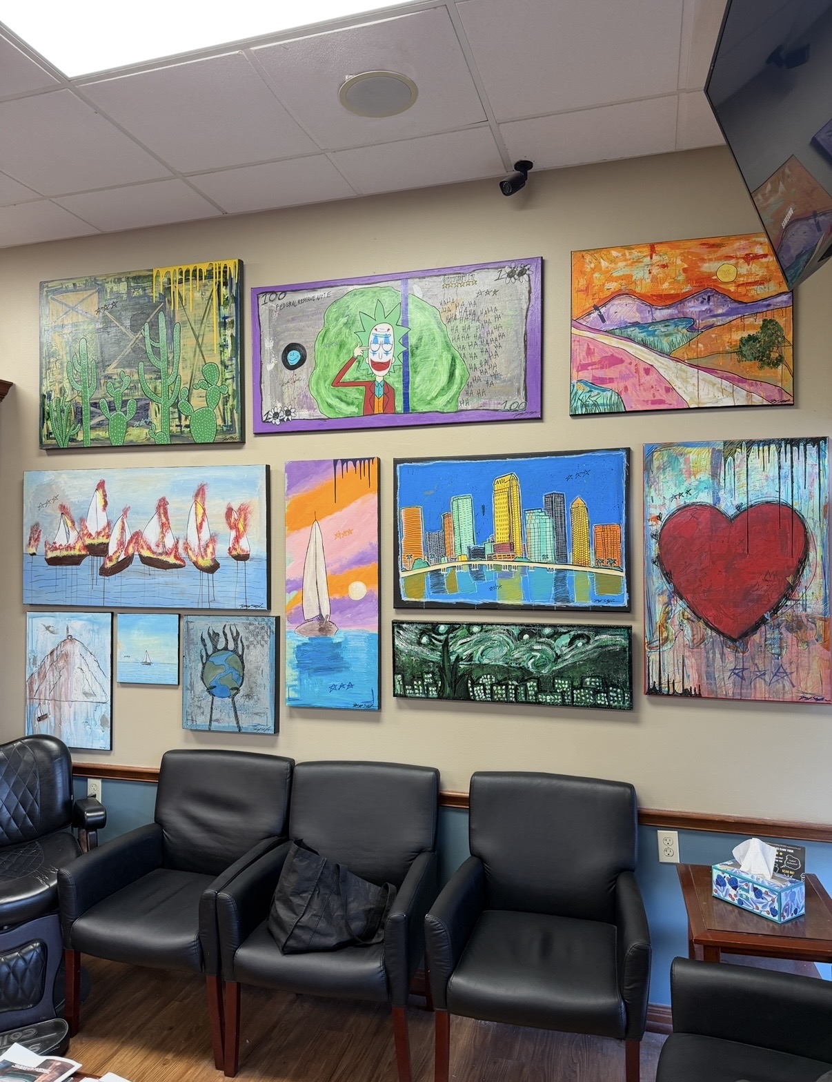 Wall with colorful paintings, black chairs, and a small table with tissue box in a room.