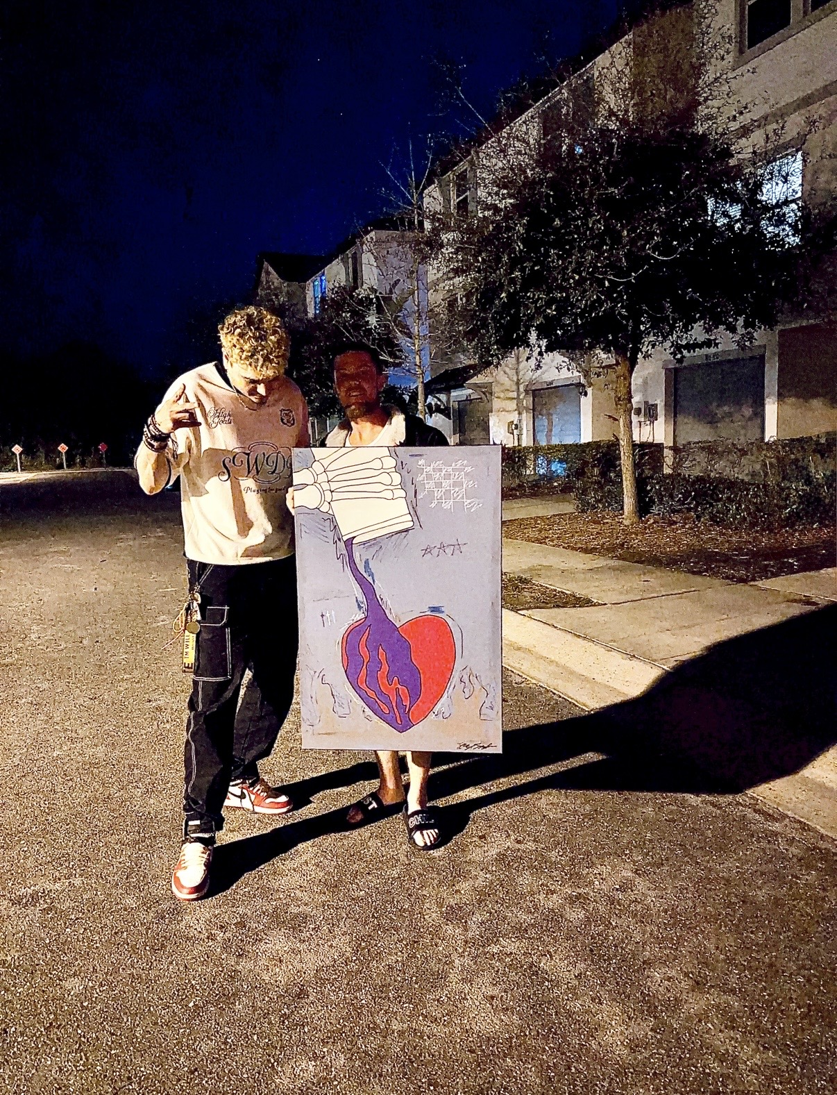 Two people holding a large painting outdoors at night, with a building and tree in the background.
