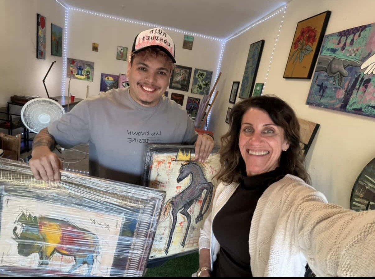 Two women smiling in an art gallery, holding a framed painting of a horse, surrounded by colorful artwork on walls.