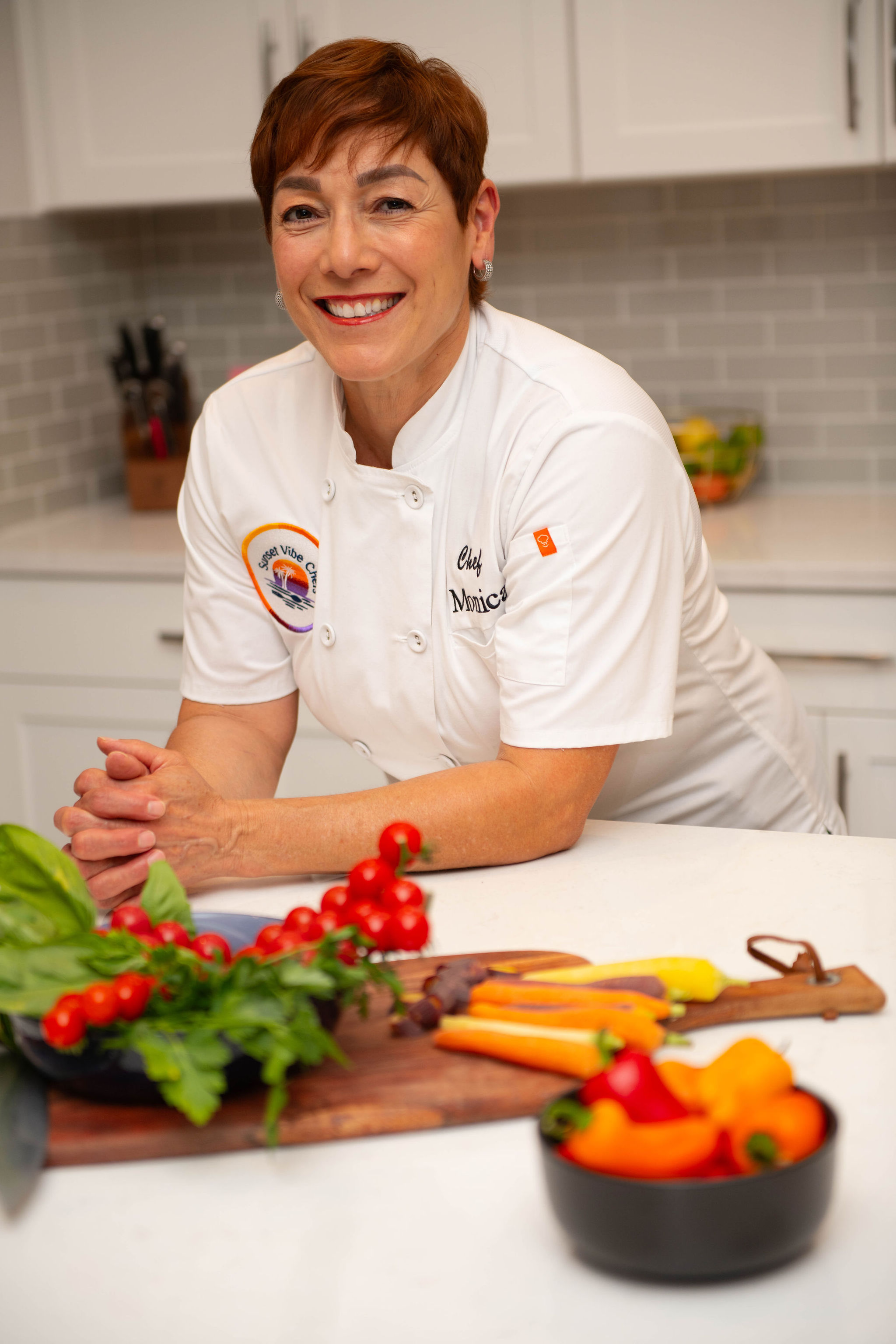 Smiling woman in chef's uniform in a kitchen with vegetables and herbs on a cutting board.