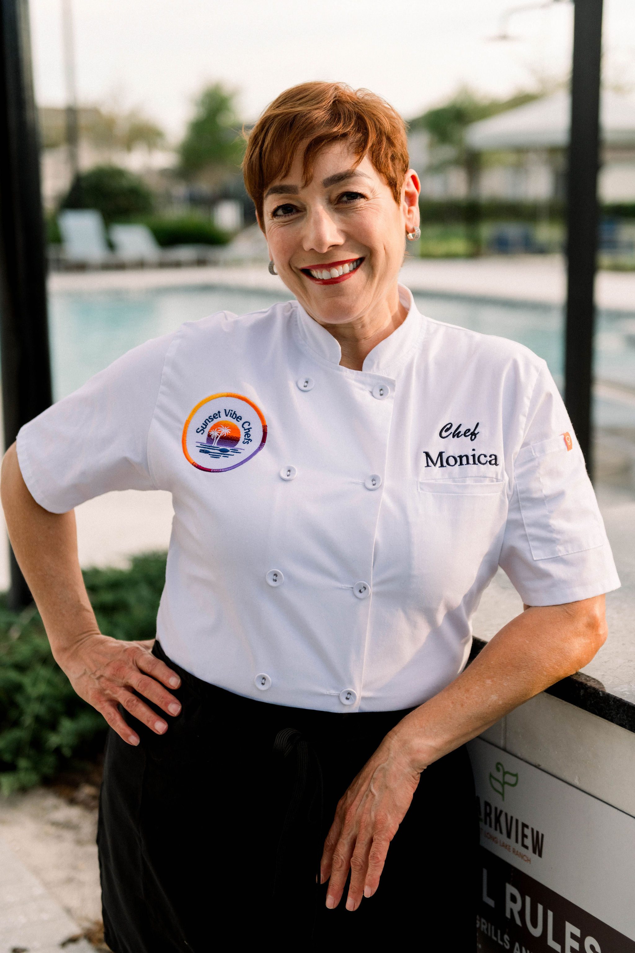 Woman in a white chef's coat with embroidered name and logo, smiling outdoors near a pool.