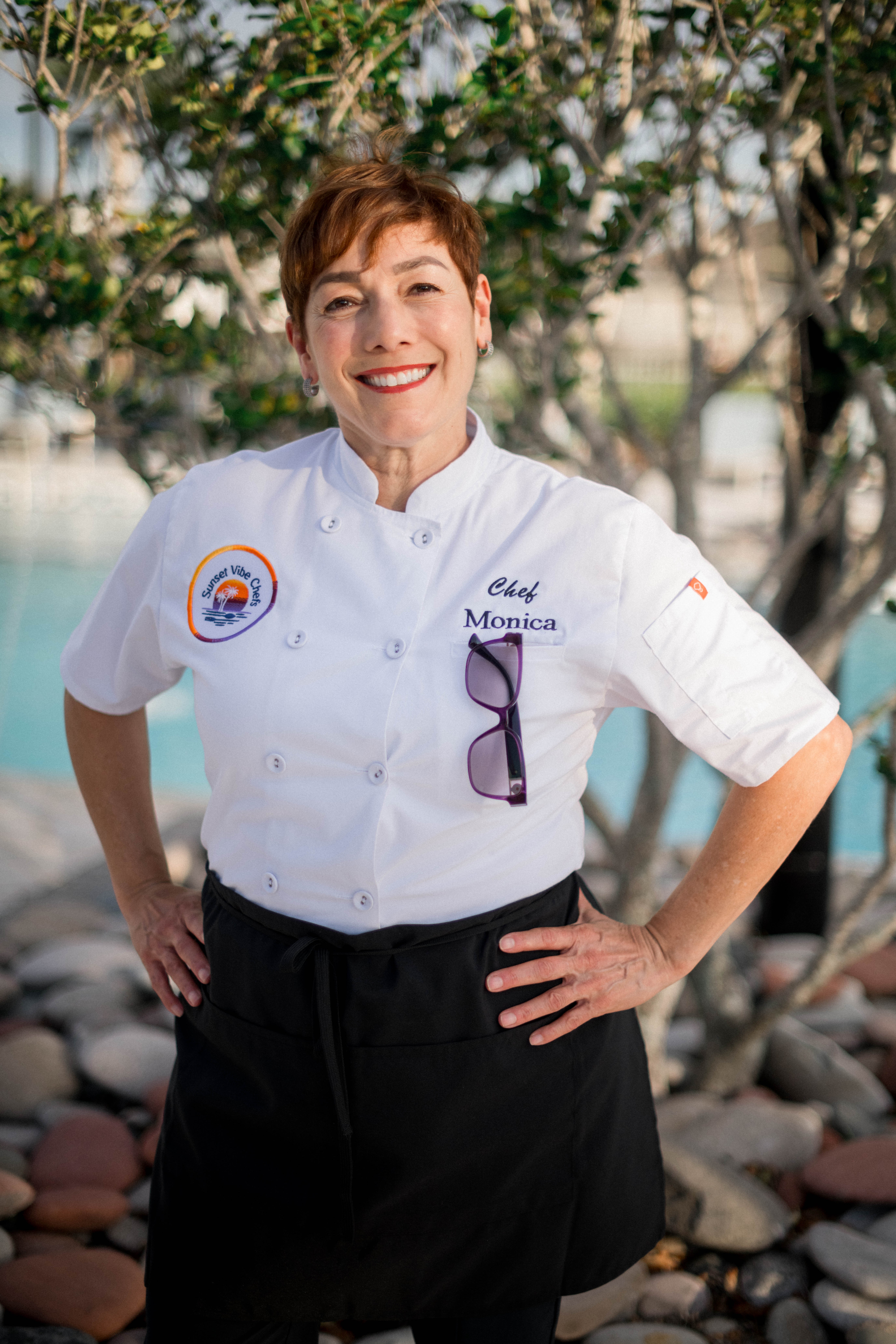 Woman in chef uniform smiling outdoors with trees and rocks in background.