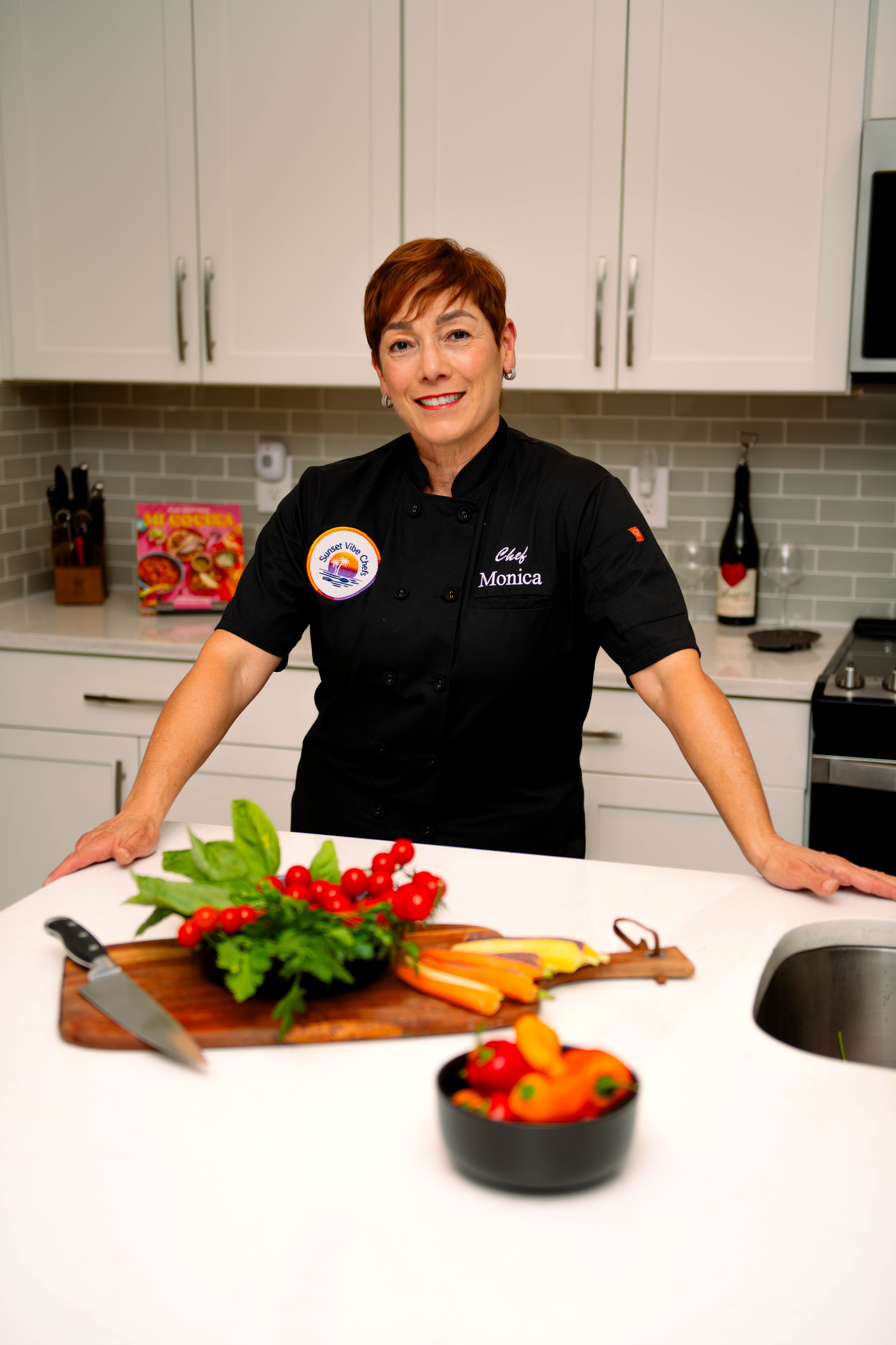 Woman in black chef's coat smiling in kitchen with vegetables and utensils on counter.