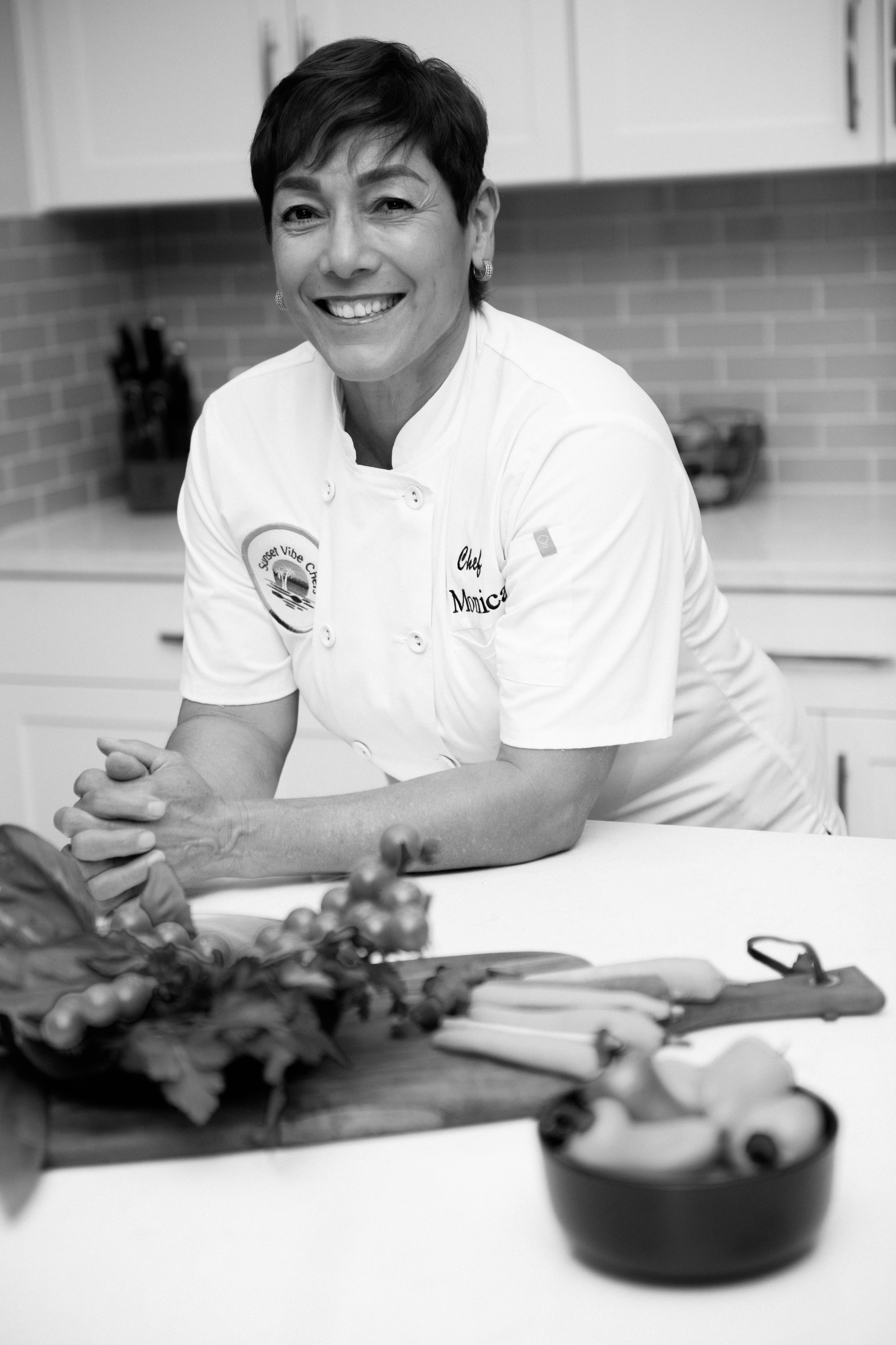 Smiling woman in chef's uniform in a kitchen with vegetables and utensils on counter.