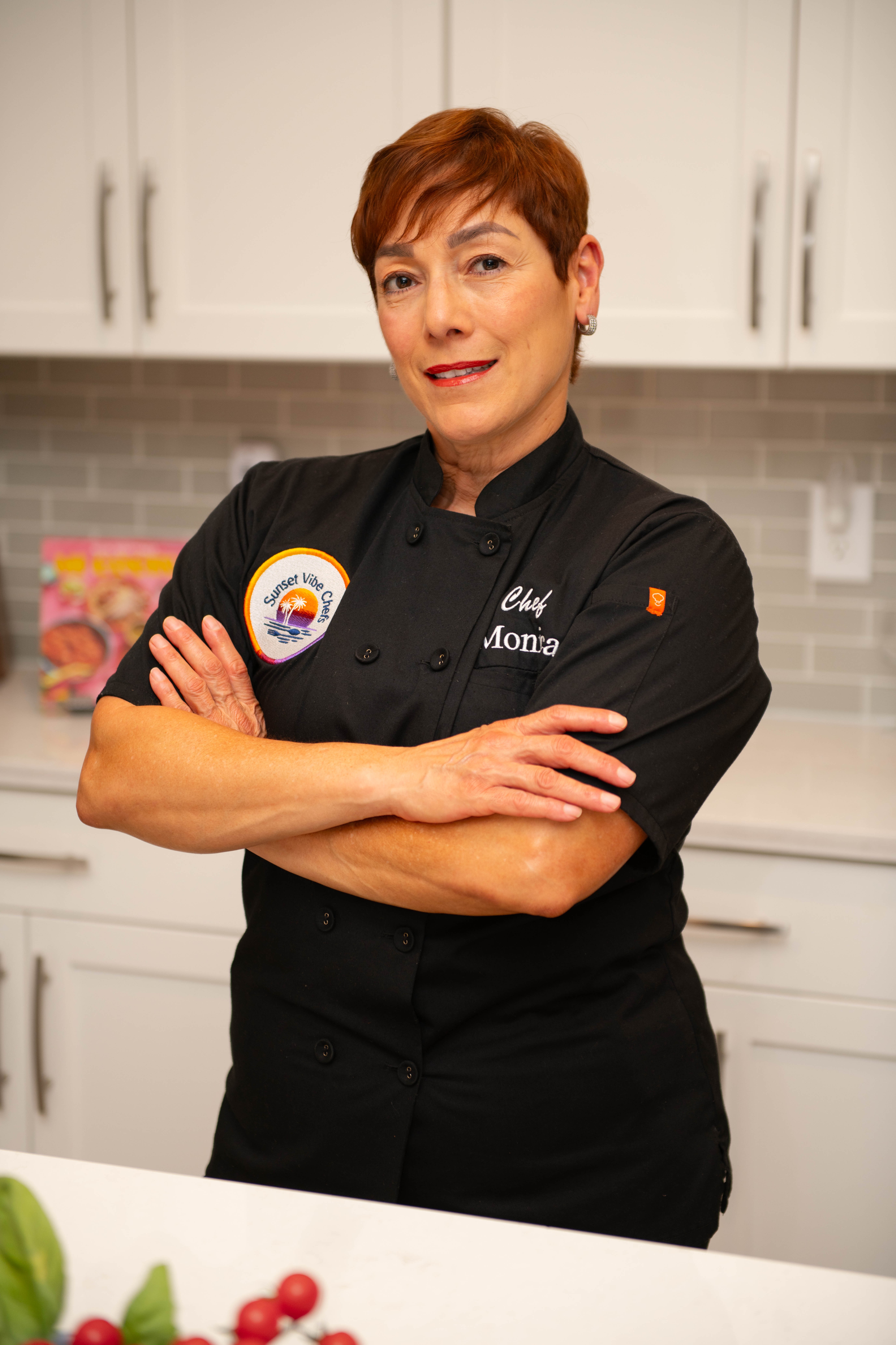 Woman in chef uniform with arms crossed, standing in a kitchen with white cabinets and gray backsplash.
