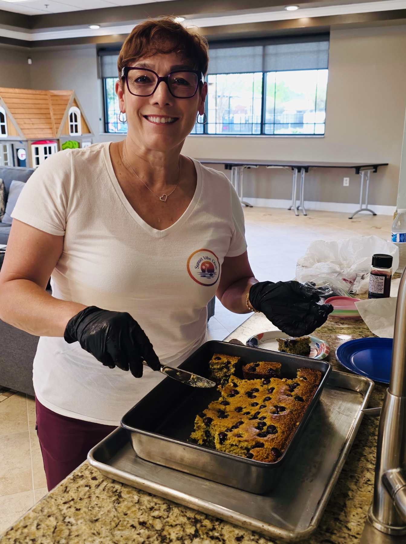 Woman wearing glasses and gloves, smiling, preparing food in a kitchen with a baking tray of baked items.