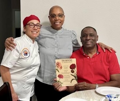 Three people standing together, one holding a book, smiling, in a room with a table and plates.