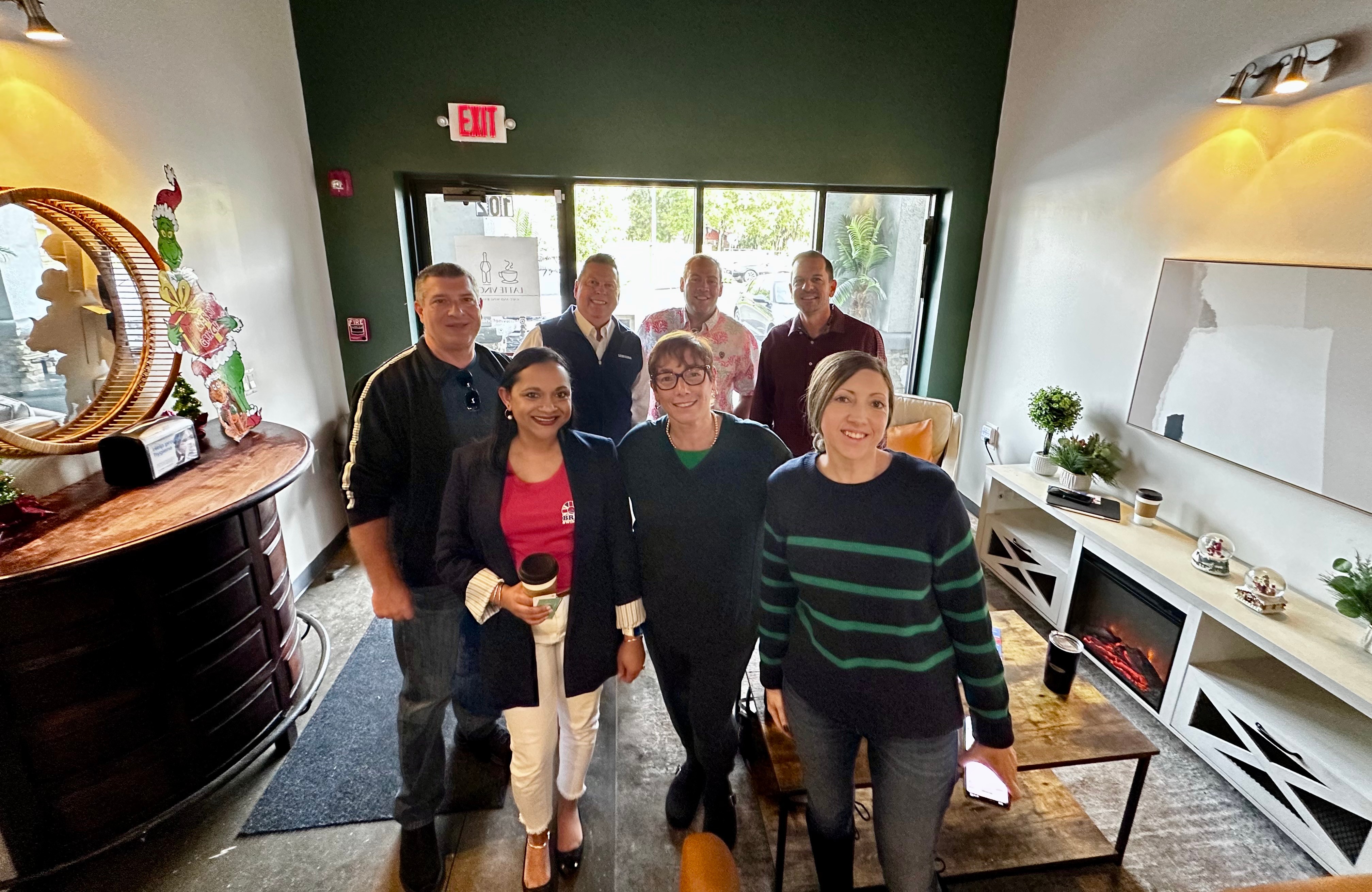 Group of seven people standing inside a room near a window, smiling and posing for the photo.