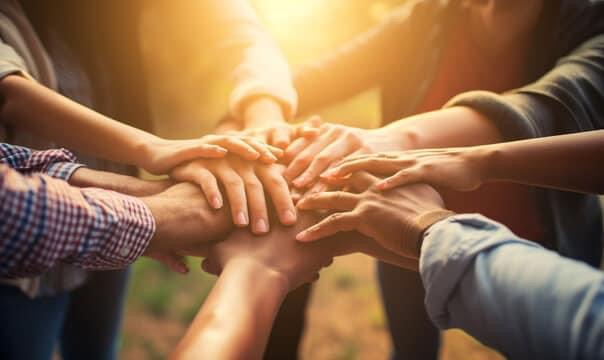 Multiple hands stacked together in a circle, symbolizing unity and teamwork, with warm sunlight in the background.