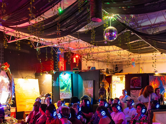 Crowd of people wearing headphones at a colorful, decorated event with screens and disco ball overhead.
