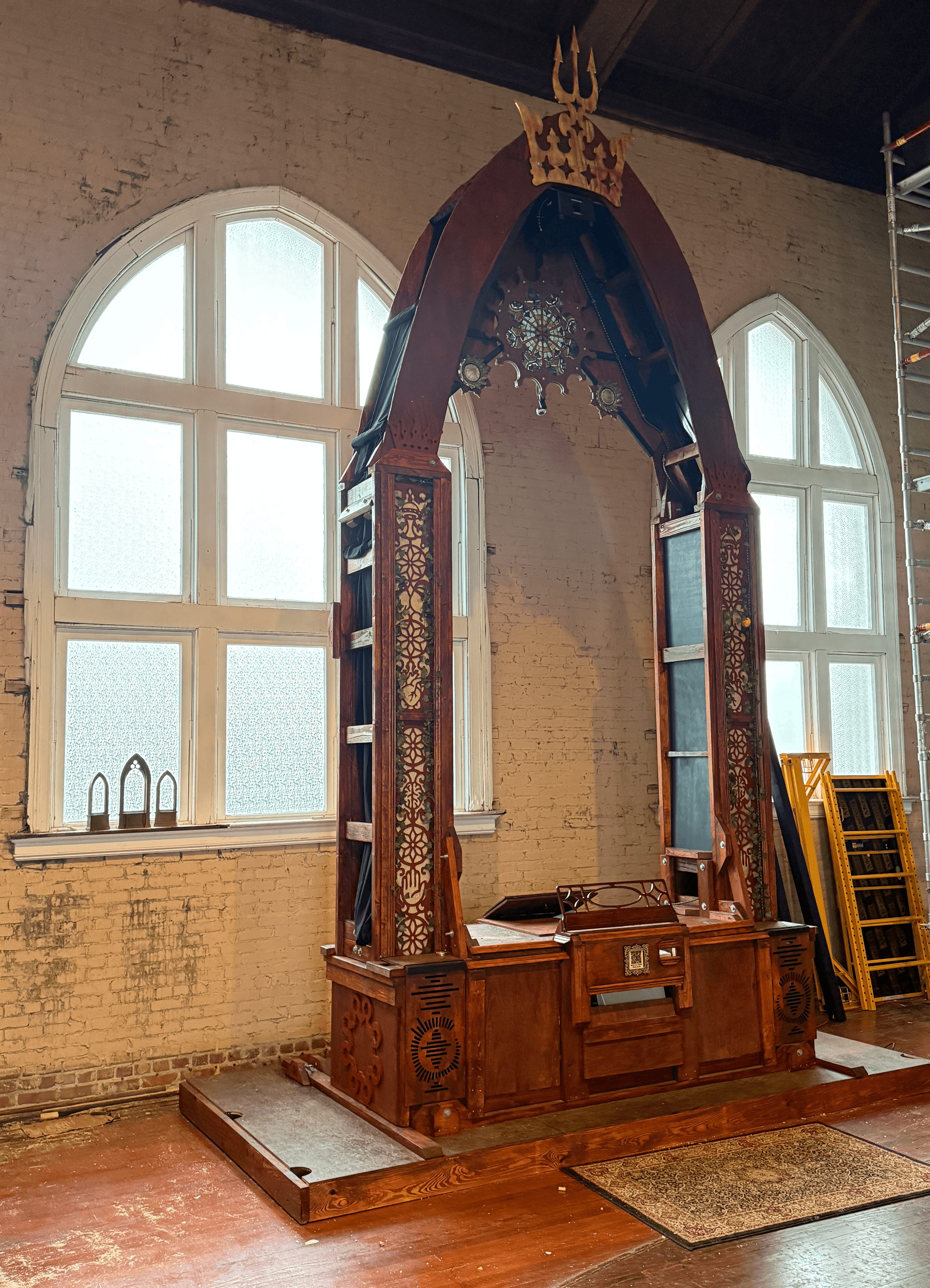 Ornate wooden structure with a crown on top, positioned between two large arched windows in a room with brick walls.