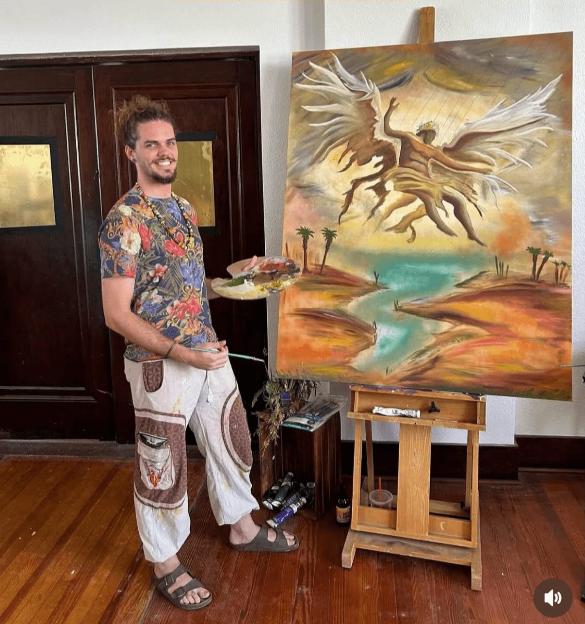 Man standing next to a colorful fantasy landscape painting on an easel, holding a palette and brush, smiling.