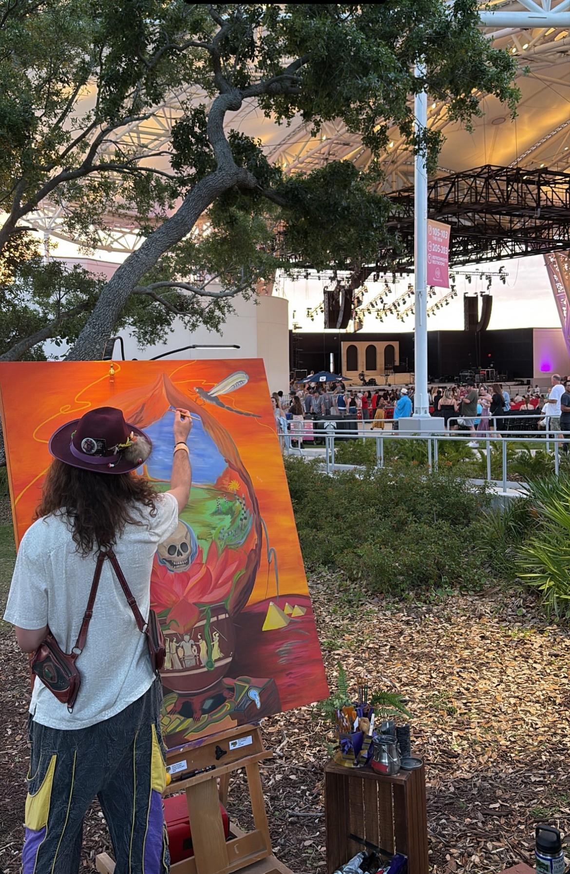 Person with hat painting on large outdoor canvas, trees and stage in background.