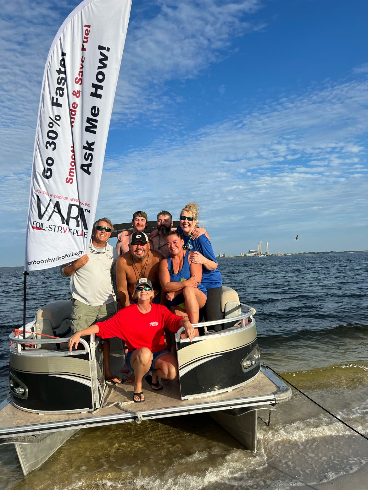 Group of seven people on a boat in water with blue sky and clouds, some wearing sunglasses, smiling.