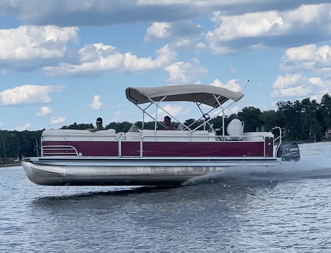 Pontoon boat with canopy moving on water, two people onboard, trees in background, partly cloudy sky.