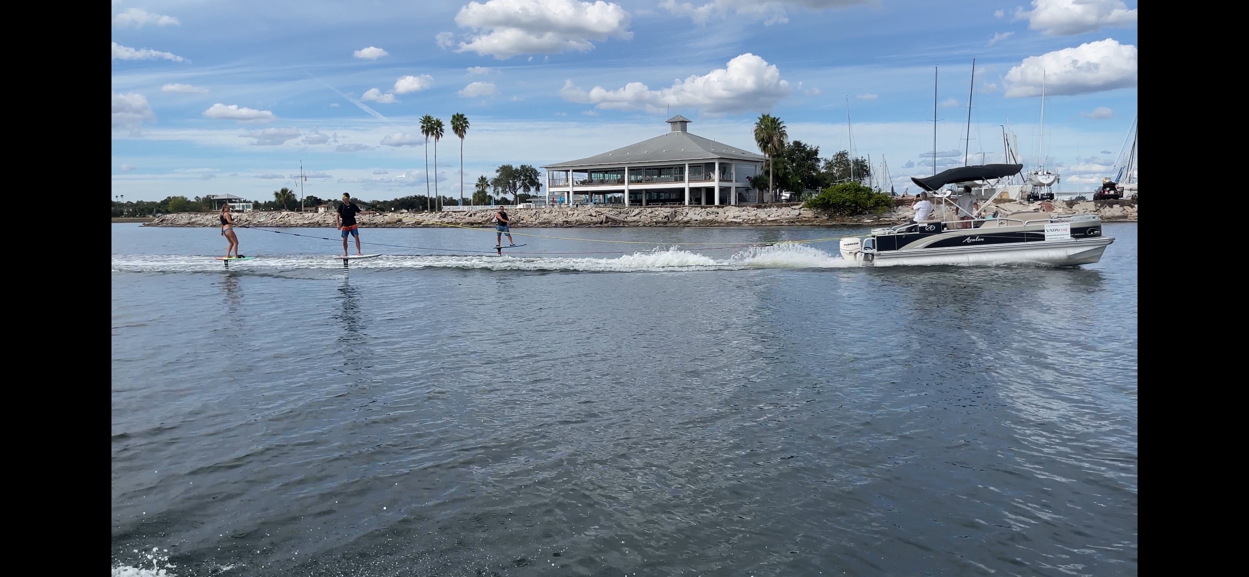 Four people walk on a dock with a boat nearby, water and sky with clouds in the background.