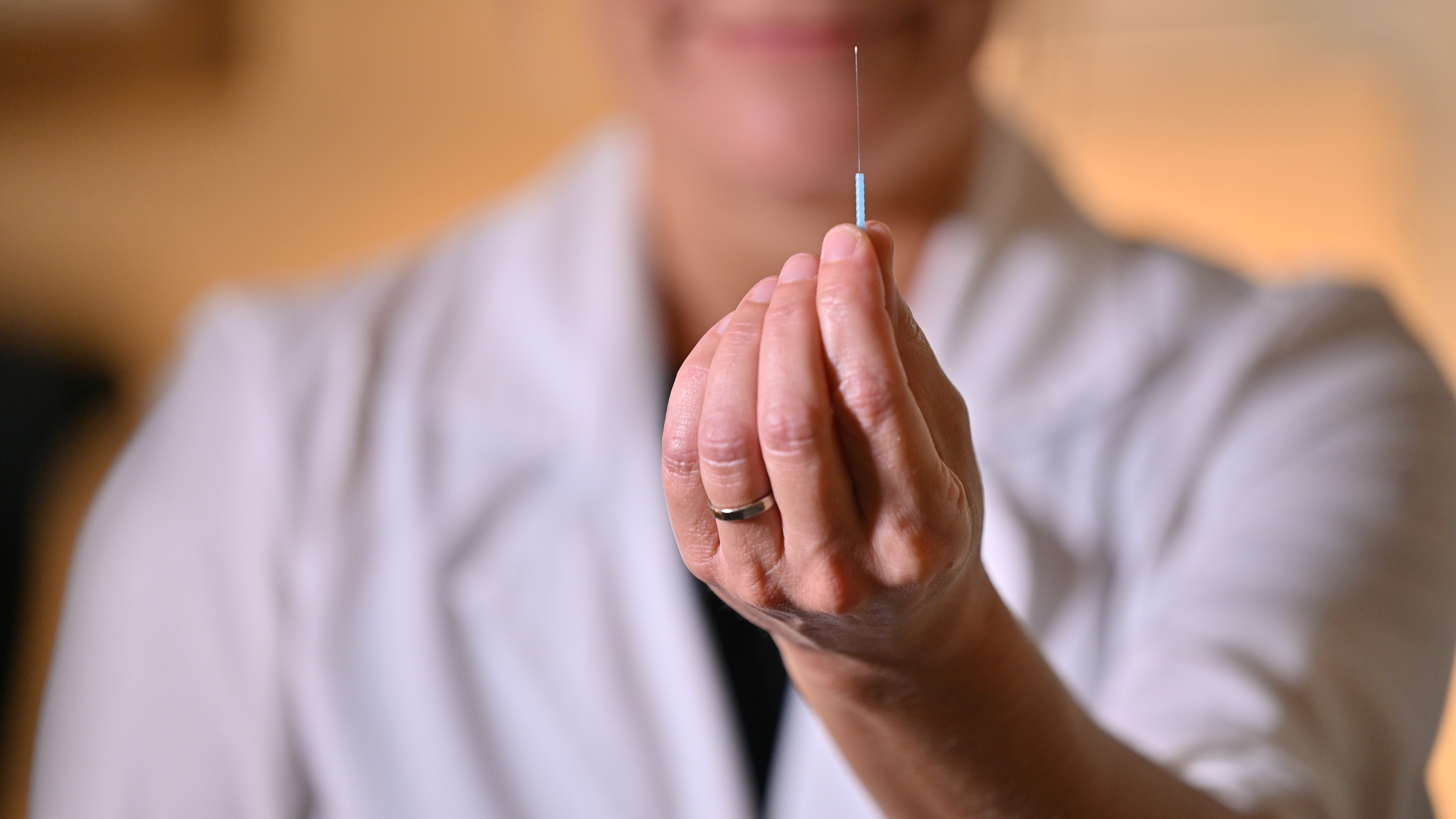Person holding a syringe close-up, focus on hand and syringe, blurred background, wearing white coat.