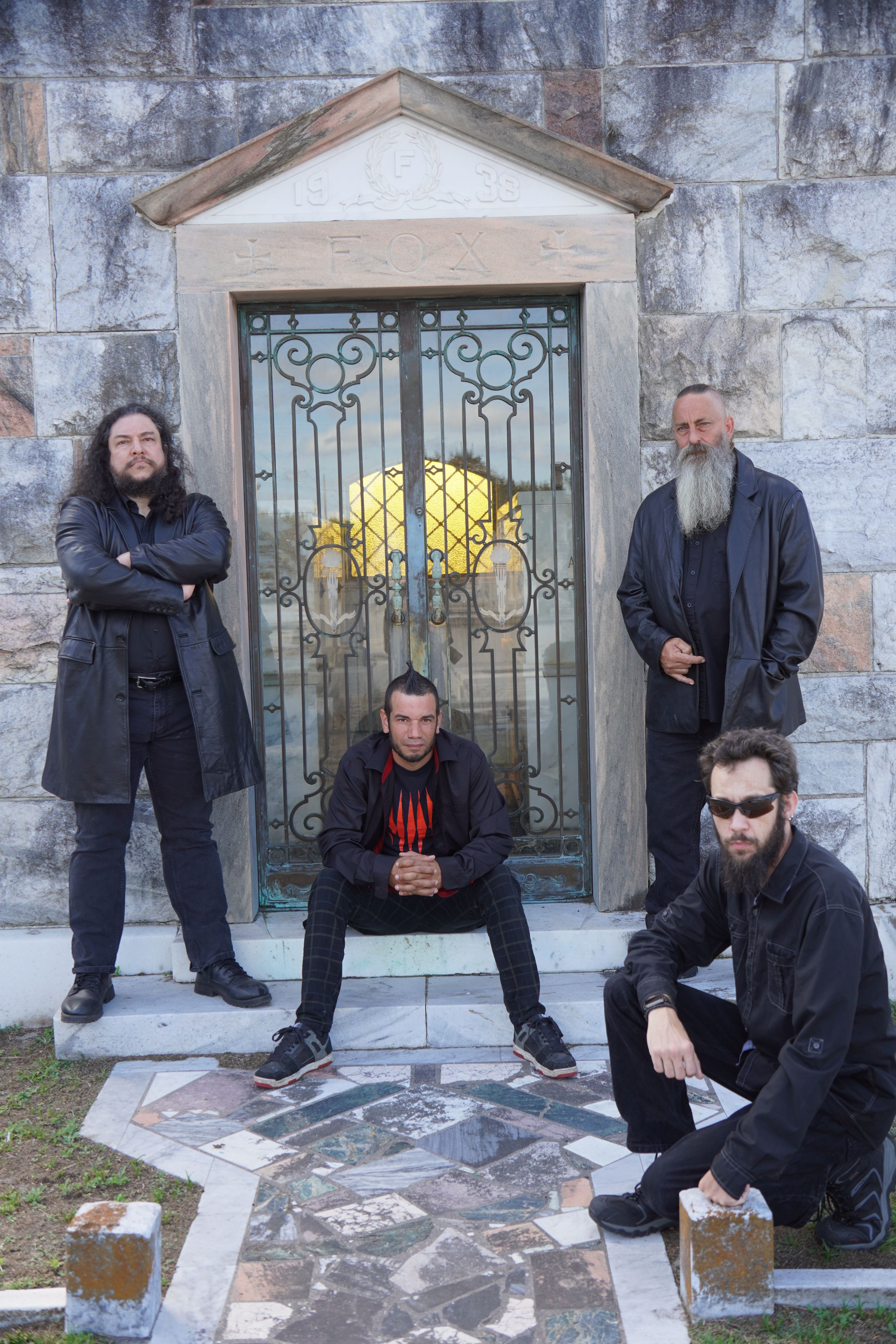 Four men with beards and dark clothing pose in front of a decorative gate and stone wall.