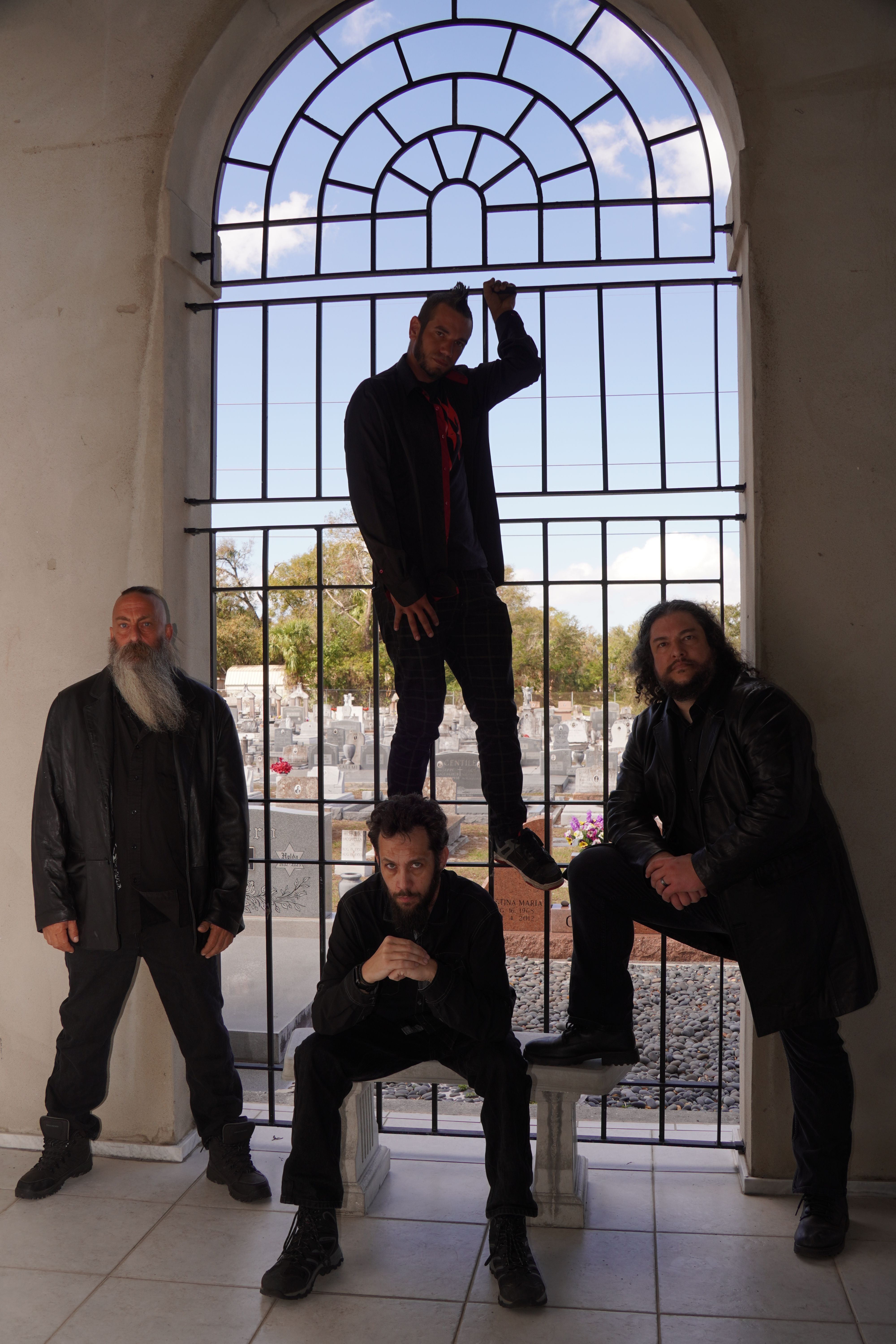 Four people pose inside a building with a large arched window and metal bars, some standing and one sitting.