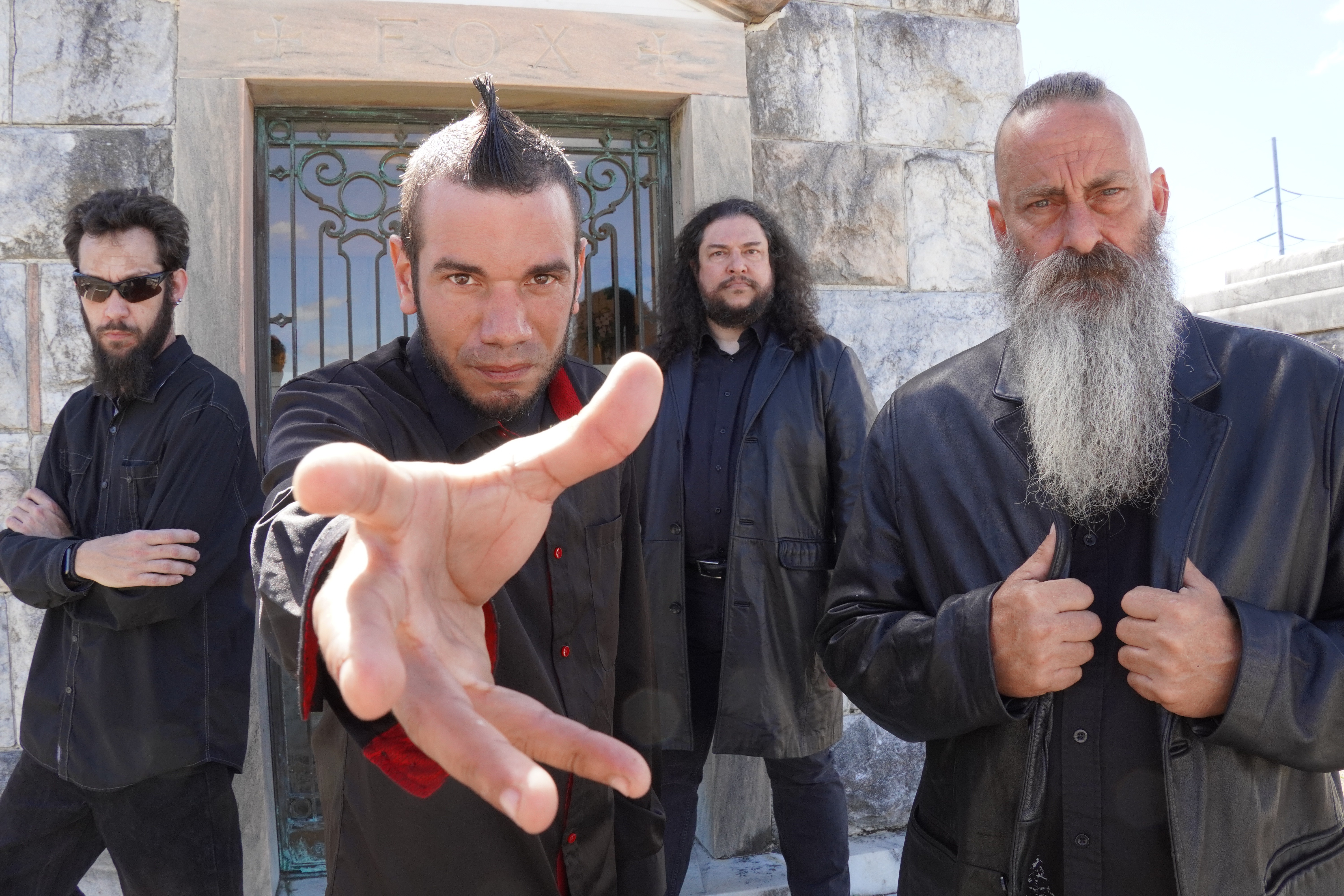 Four men standing outside a stone building with a metal gate, one reaching toward the camera.
