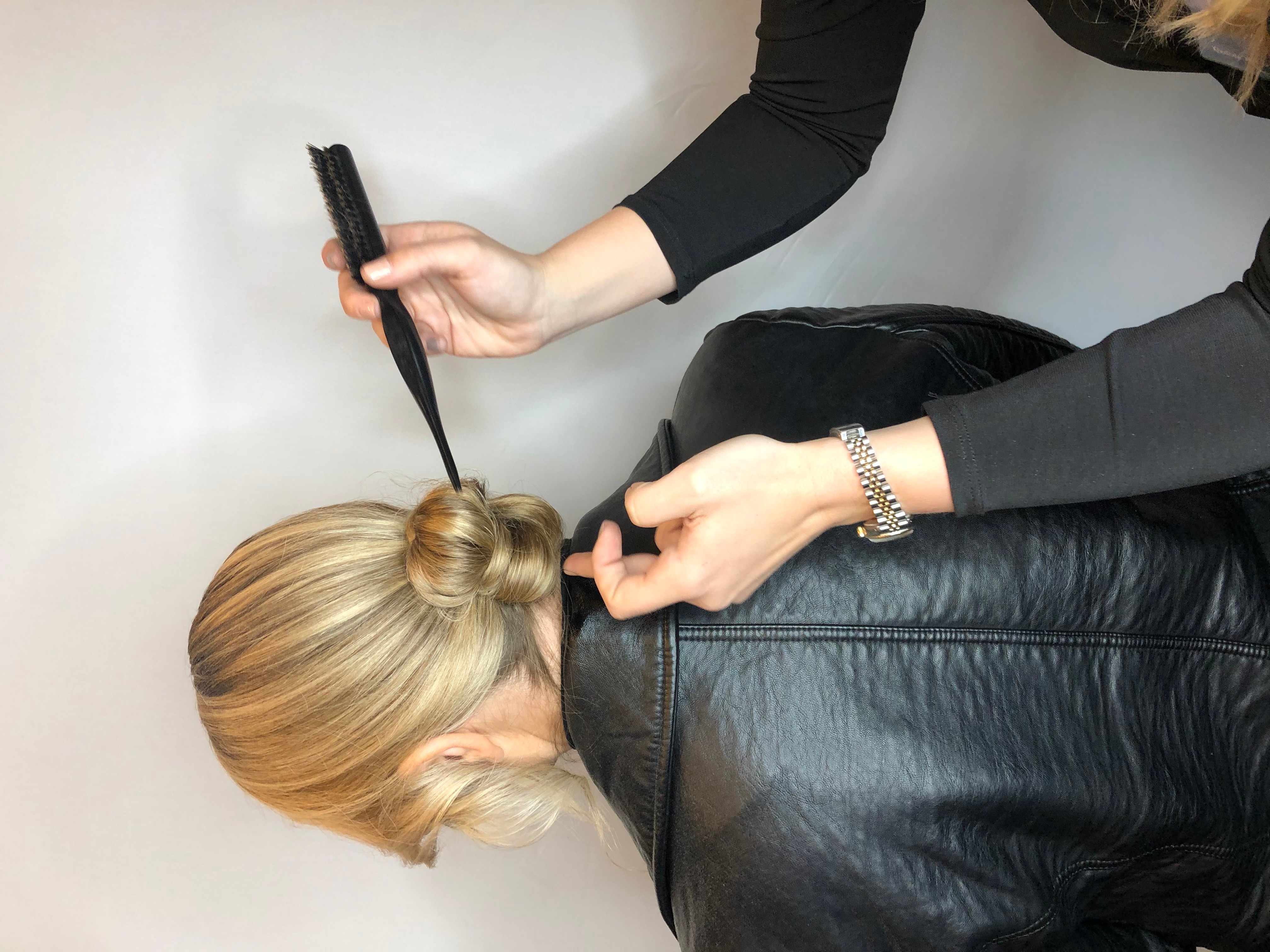 Person styling a woman's hair with a flat iron, woman wearing a black leather jacket, person wearing a black long-sleeve shirt, hair styled in a bun