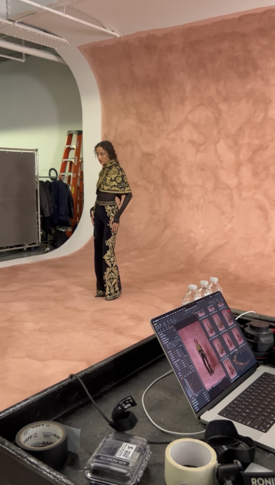 Woman standing on a pink backdrop in a studio, with a laptop and camera equipment in foreground.