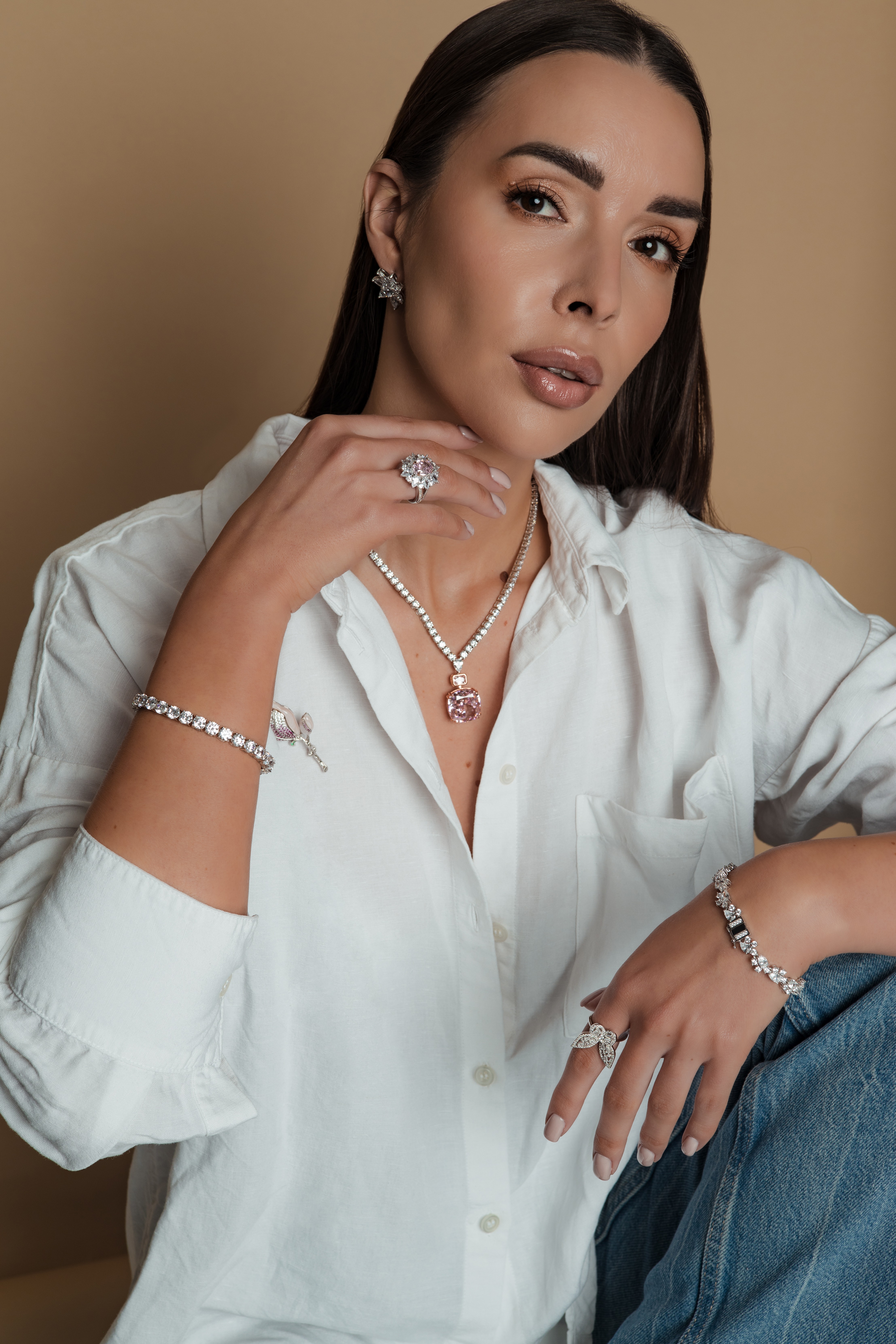 Woman with long dark hair wearing jewelry and a white shirt, sitting against a beige background.