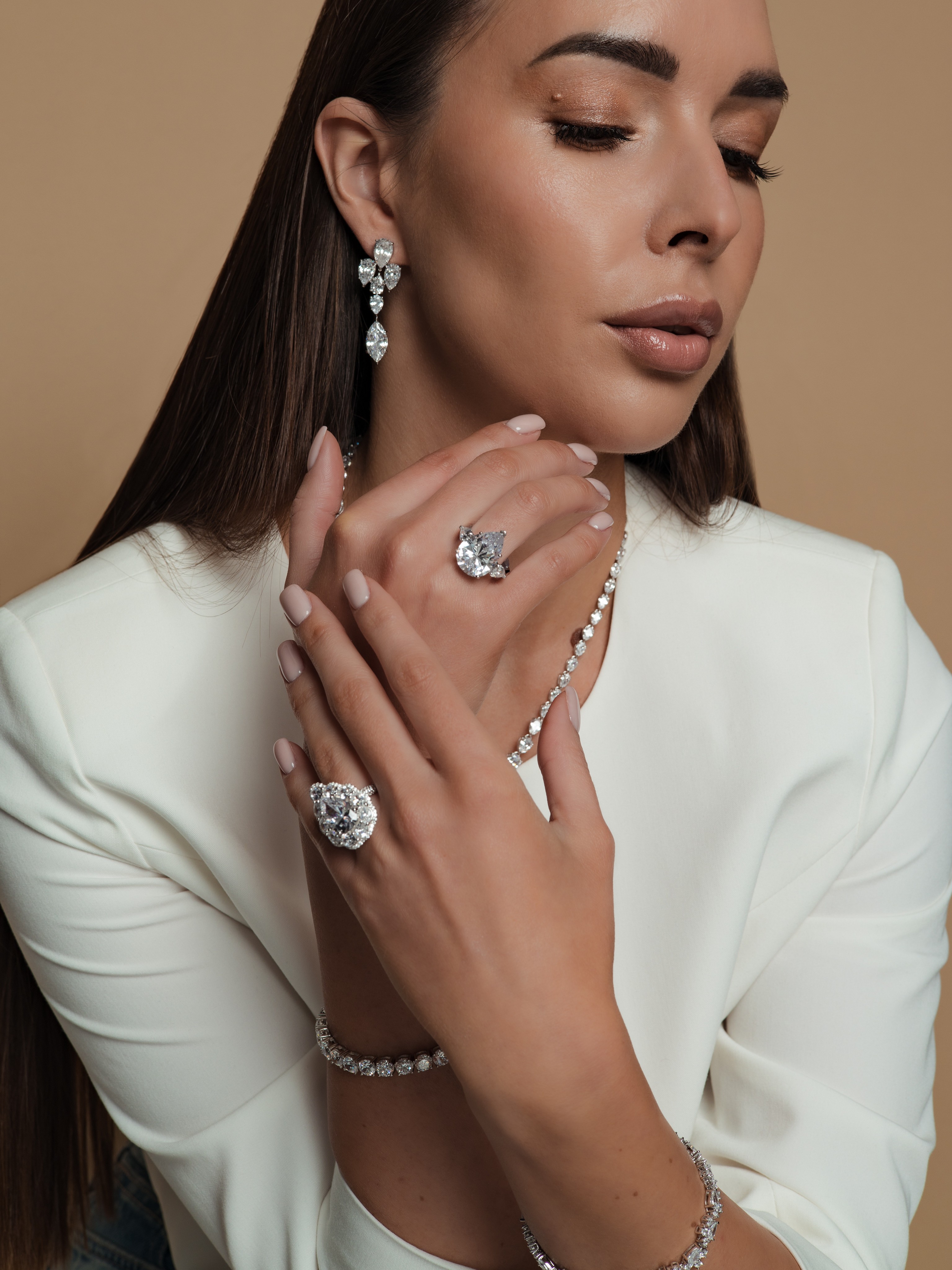 Woman with long dark hair wearing white outfit, jewelry including earrings, rings, necklace, and bracelet, posed with hands near face.