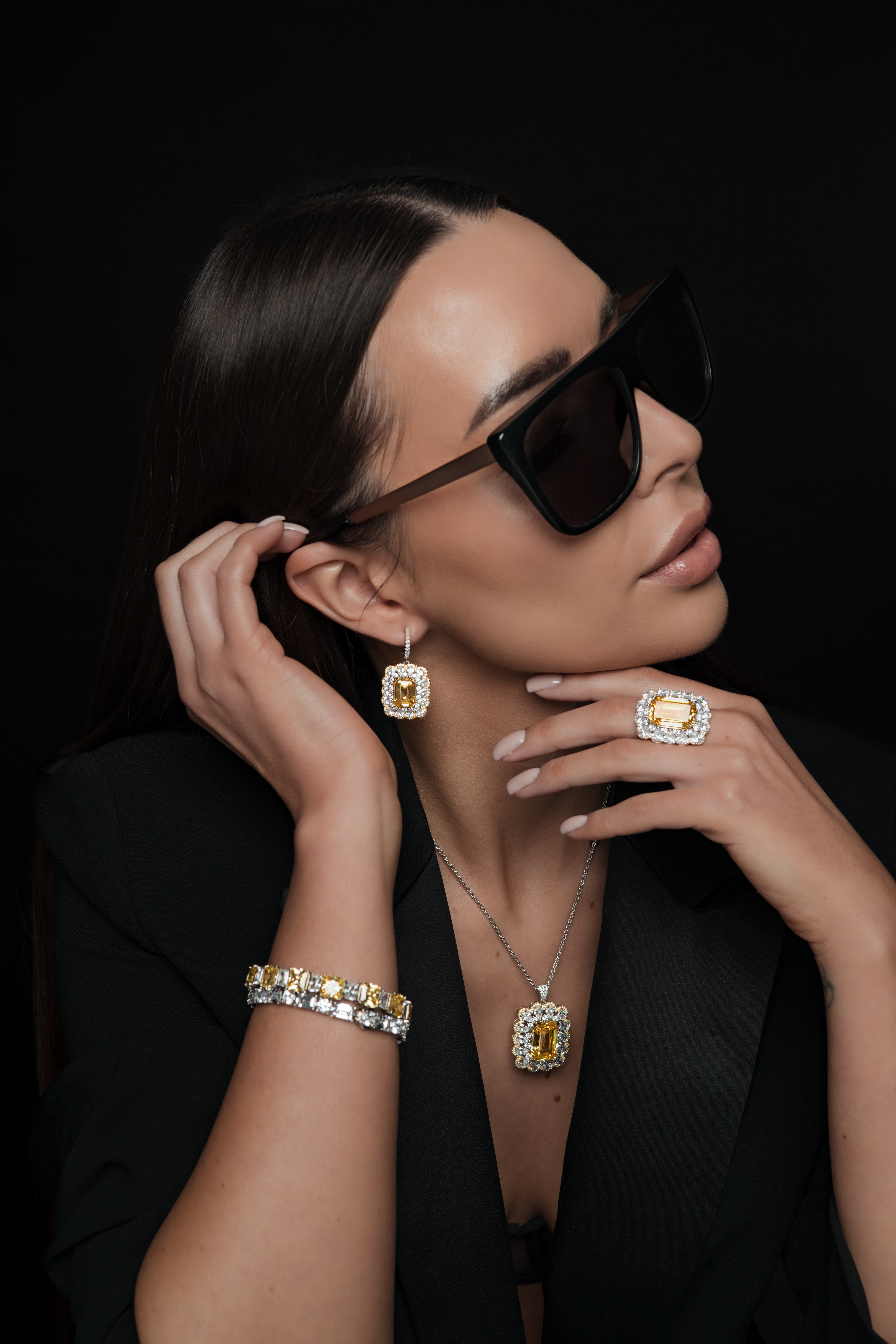 Woman with dark hair wearing large sunglasses, earrings, ring, bracelet, and necklace, posed against black background.