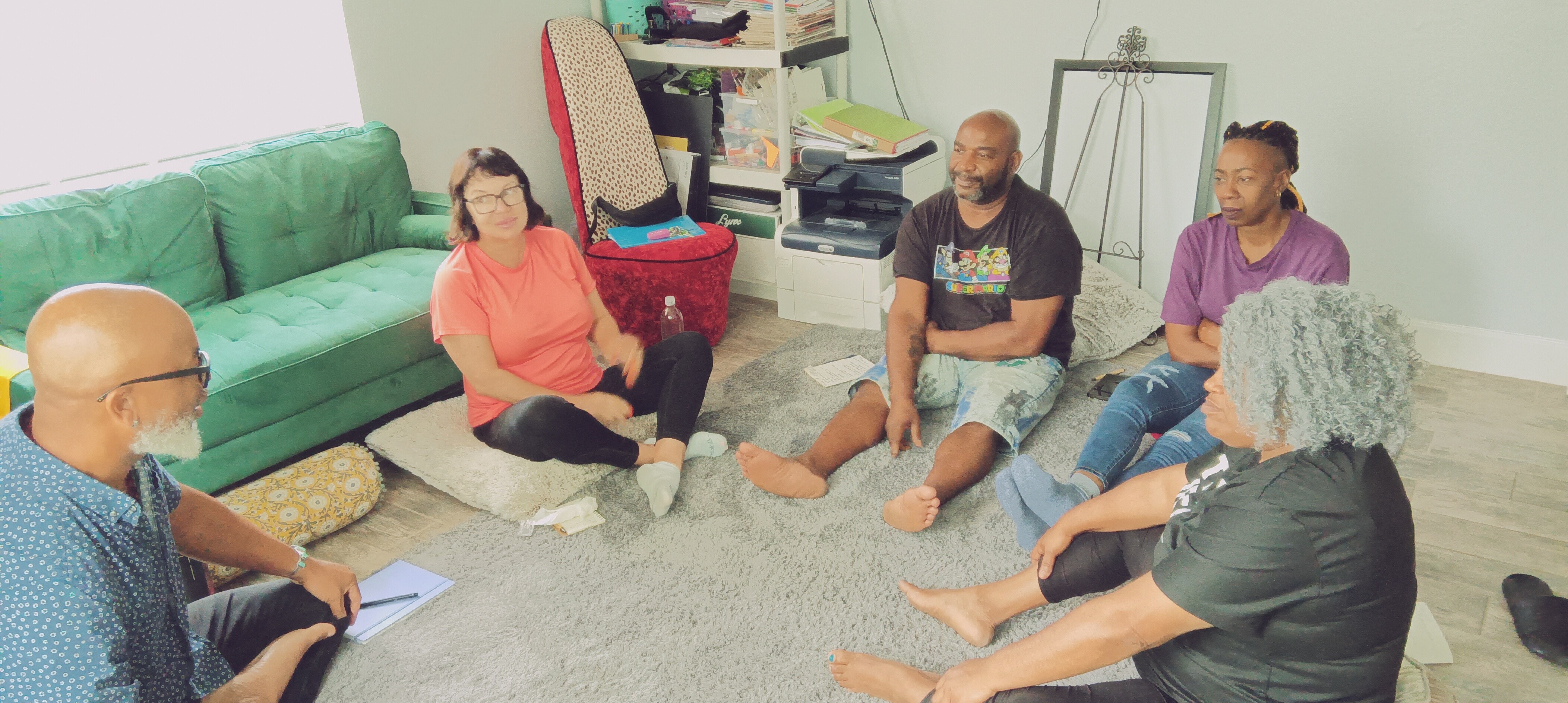 Six people sit on the floor and a sofa in a circle during a discussion in a room.